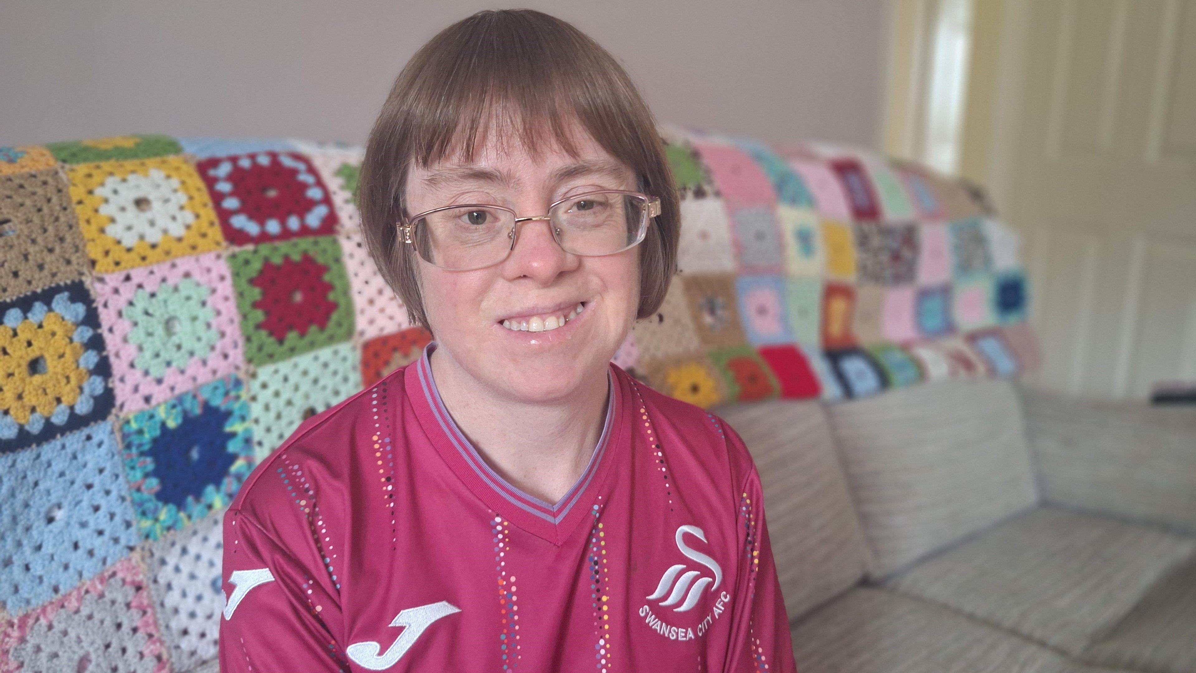 Sara Pickard is sitting on a beige sofa. The sofa has a colourful crocheted blanket thrown over the back of it. Sara is smiling and looking into camera. She has light brown hair in a bob with a fringe and wears glasses. She is wearing a burgundy Swansea City football shirt.