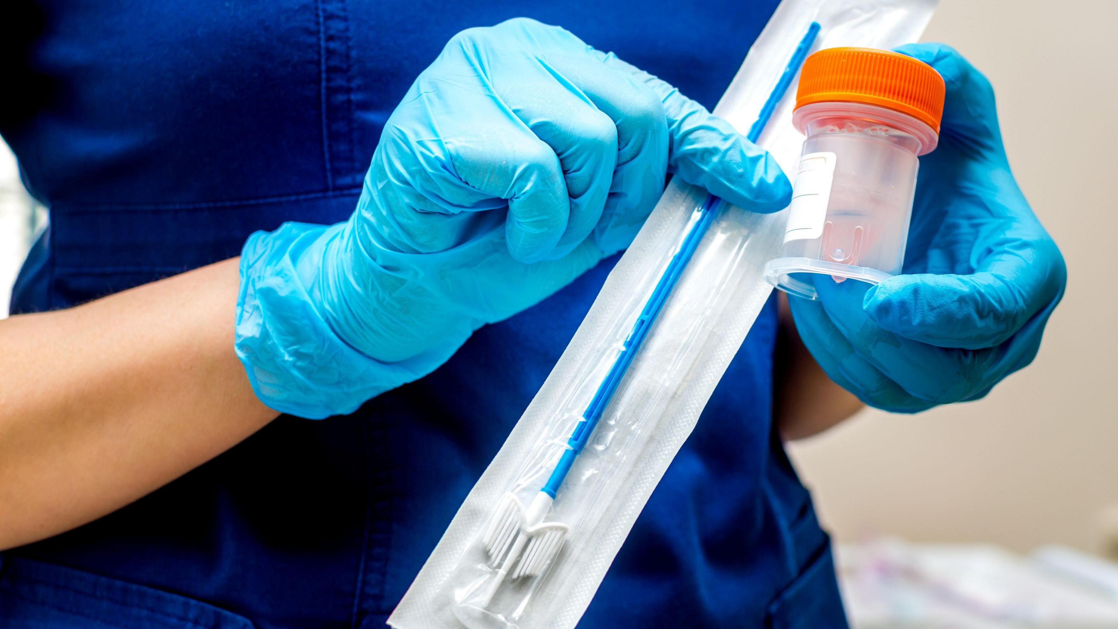 A Gynecologist, wearing blue gloves and a blue uniform, holds a brush and tube for sampling 