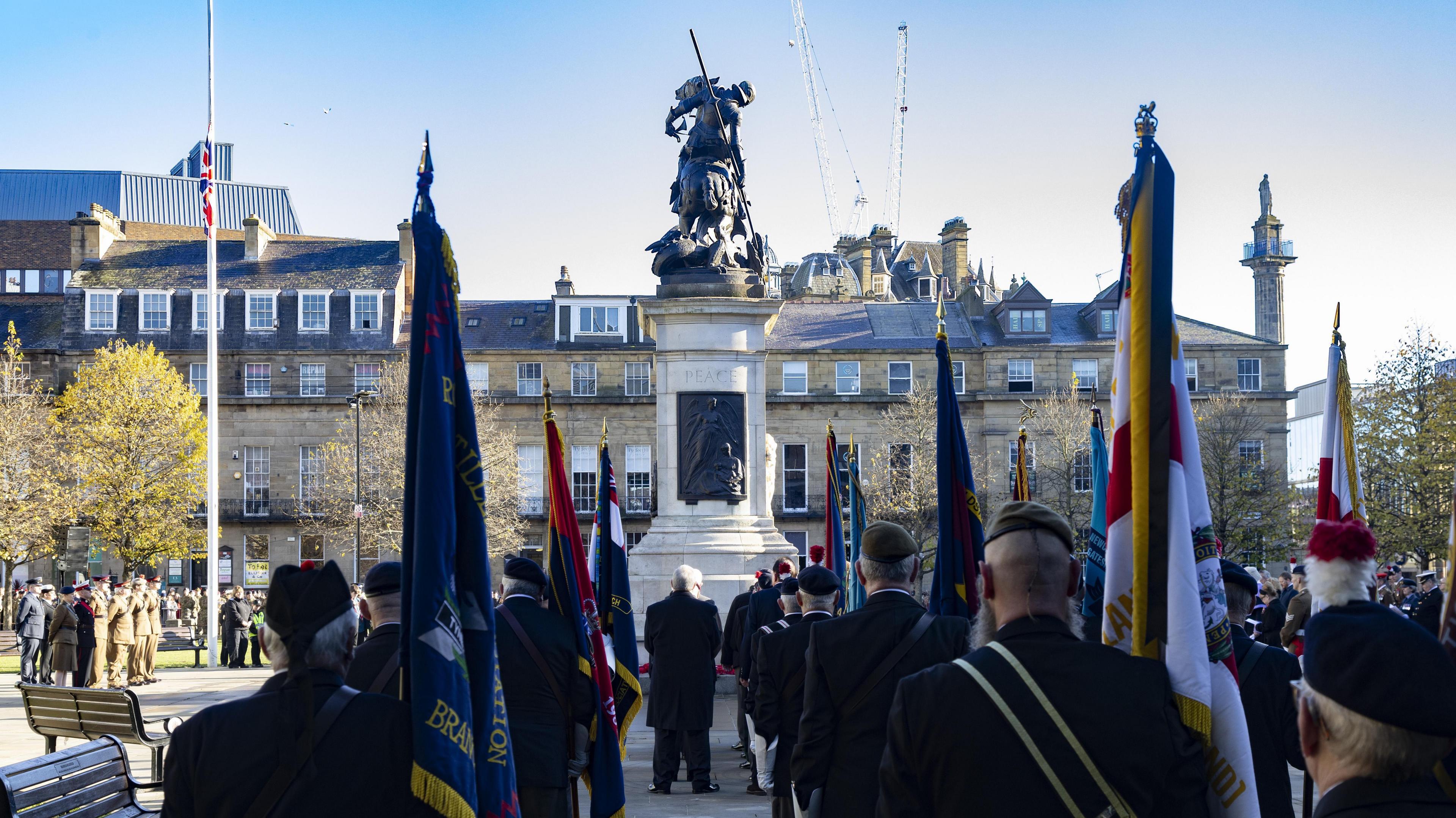 A group of veterans in uniform pictured from behind, standing facing the war memorial in Newcastle city centre. All around the monument are other groups of people standing facing the statue, which portrays Saint George slaying the dragon atop a granite stone pillar.