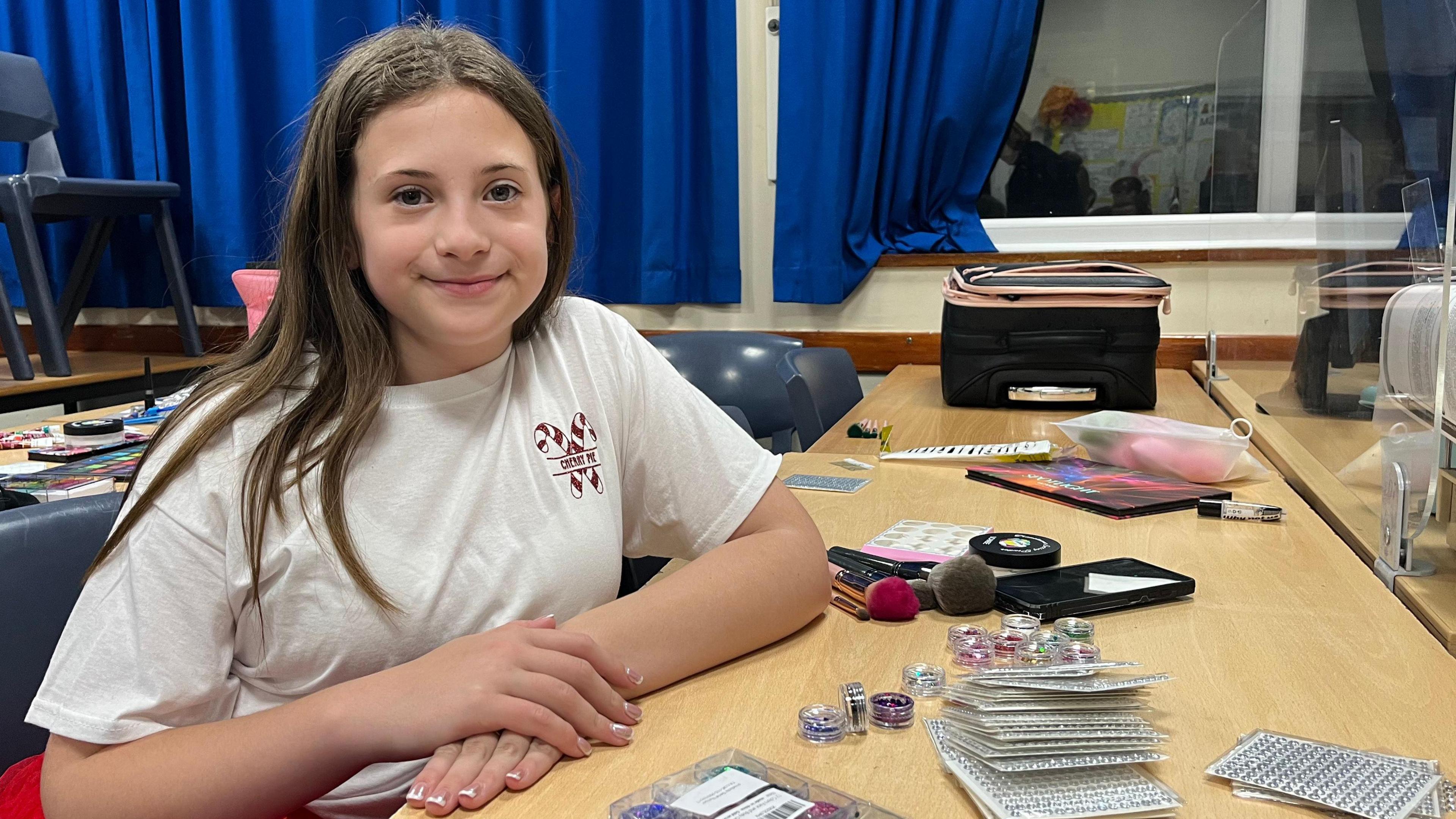 Lily sits in front of a table full of make up items. She has long brown hair and wears a white t-shirt and red skirt. 