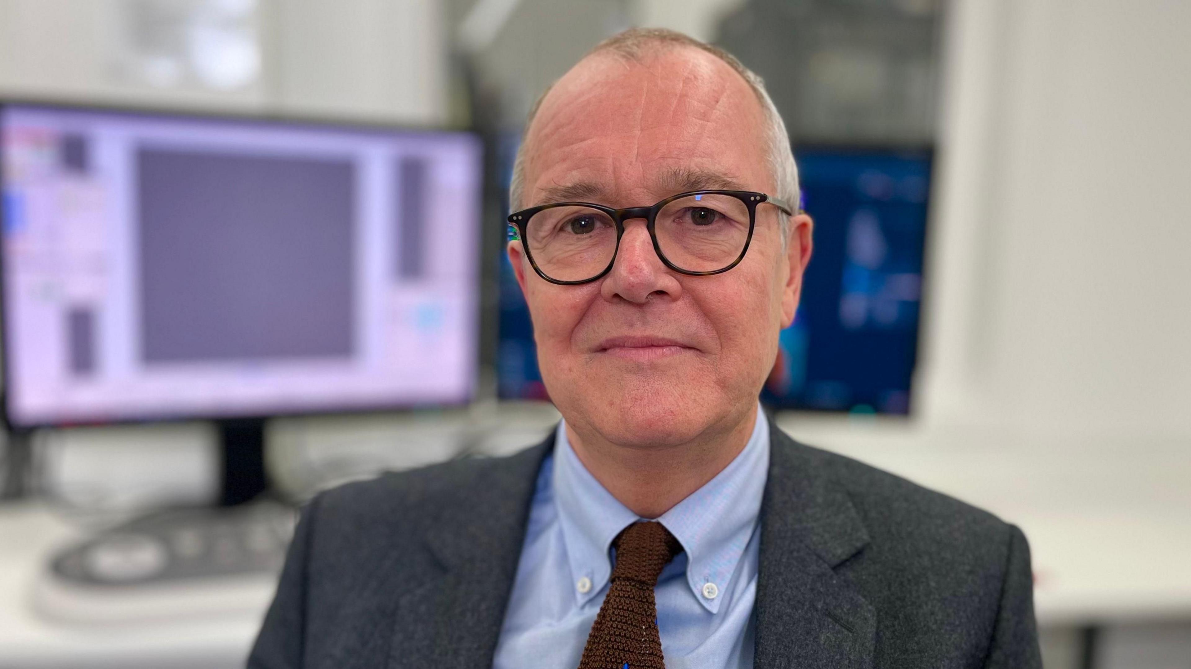 Patrick Vallance sitting in front of a desk with a computer screen. He is wearing glasses, a pale blue shirt, deep red tie and dark coloured tweed jacket.