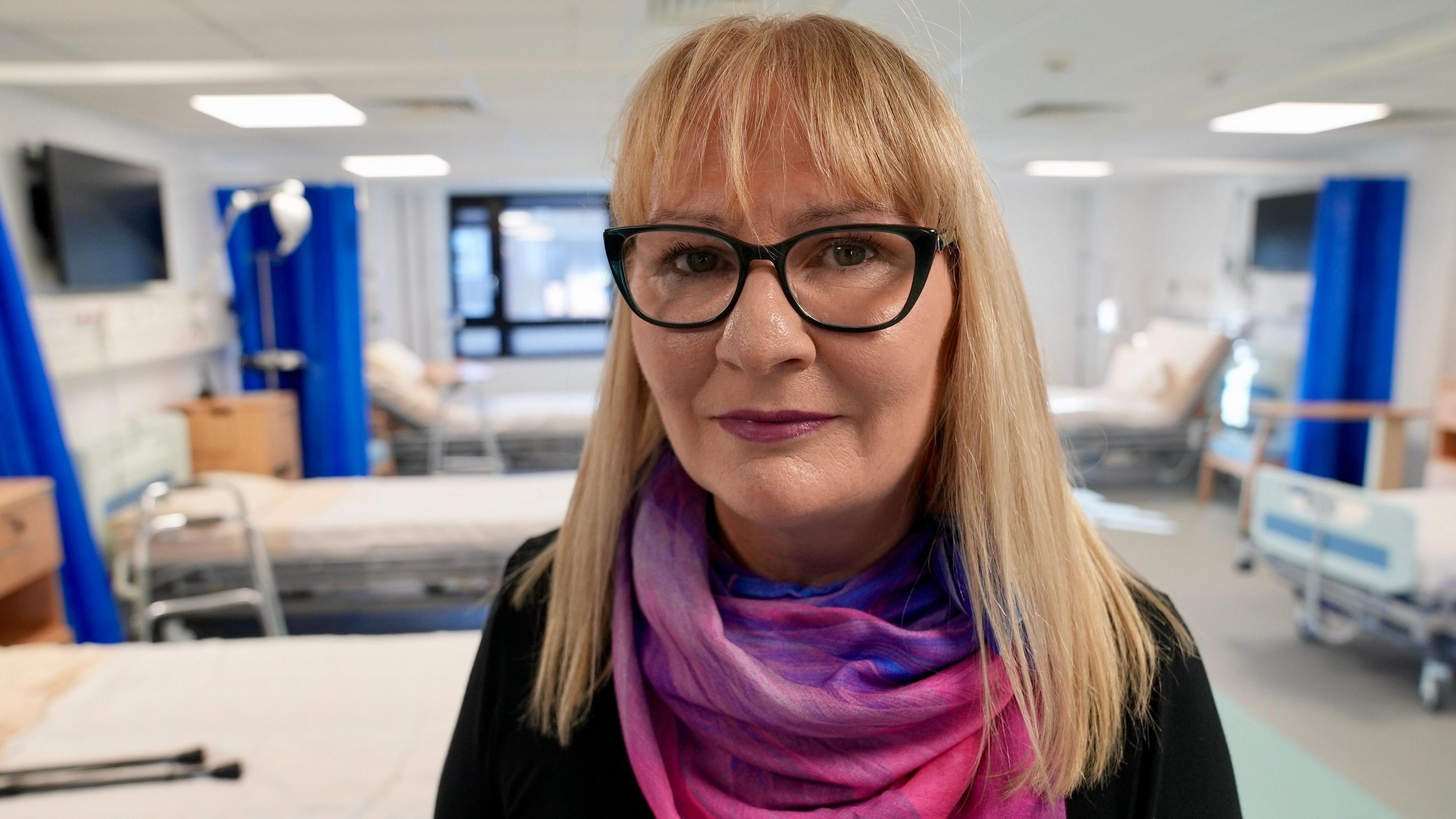 Lesley Butcher - menyw gwallt golau mewn top du, sgarff binc a sbectol, yn sefyll mewn ward ysbyty ymarfer, gyda sawl gwely gwag yn y cefndir