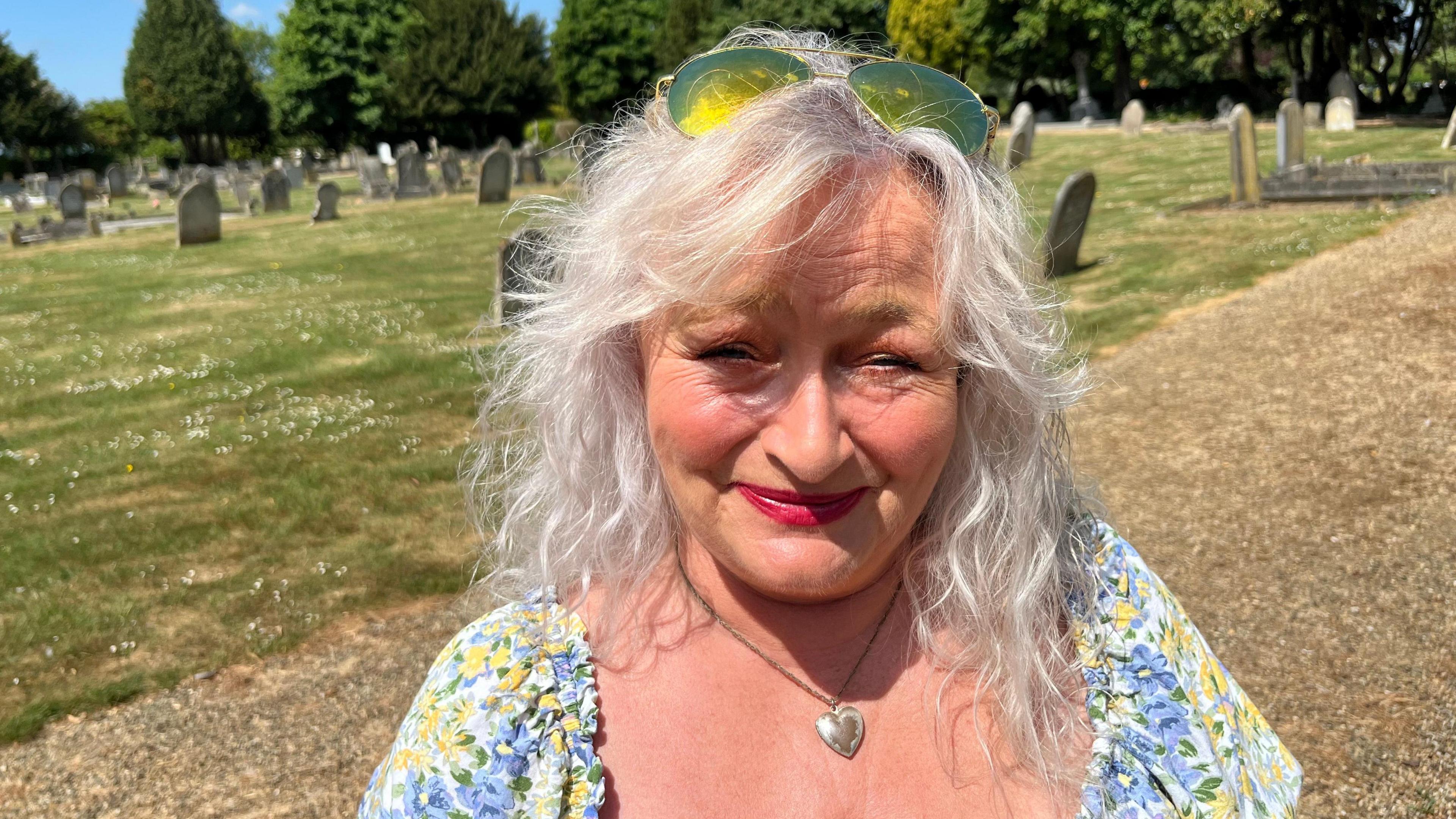 Sally Leckie, 59, has white wavy shoulder-length hair with sunglasses perched on her head. She has a floral dress with a silver heart on a necklace. Behind her are gravestones surrounded by grass.