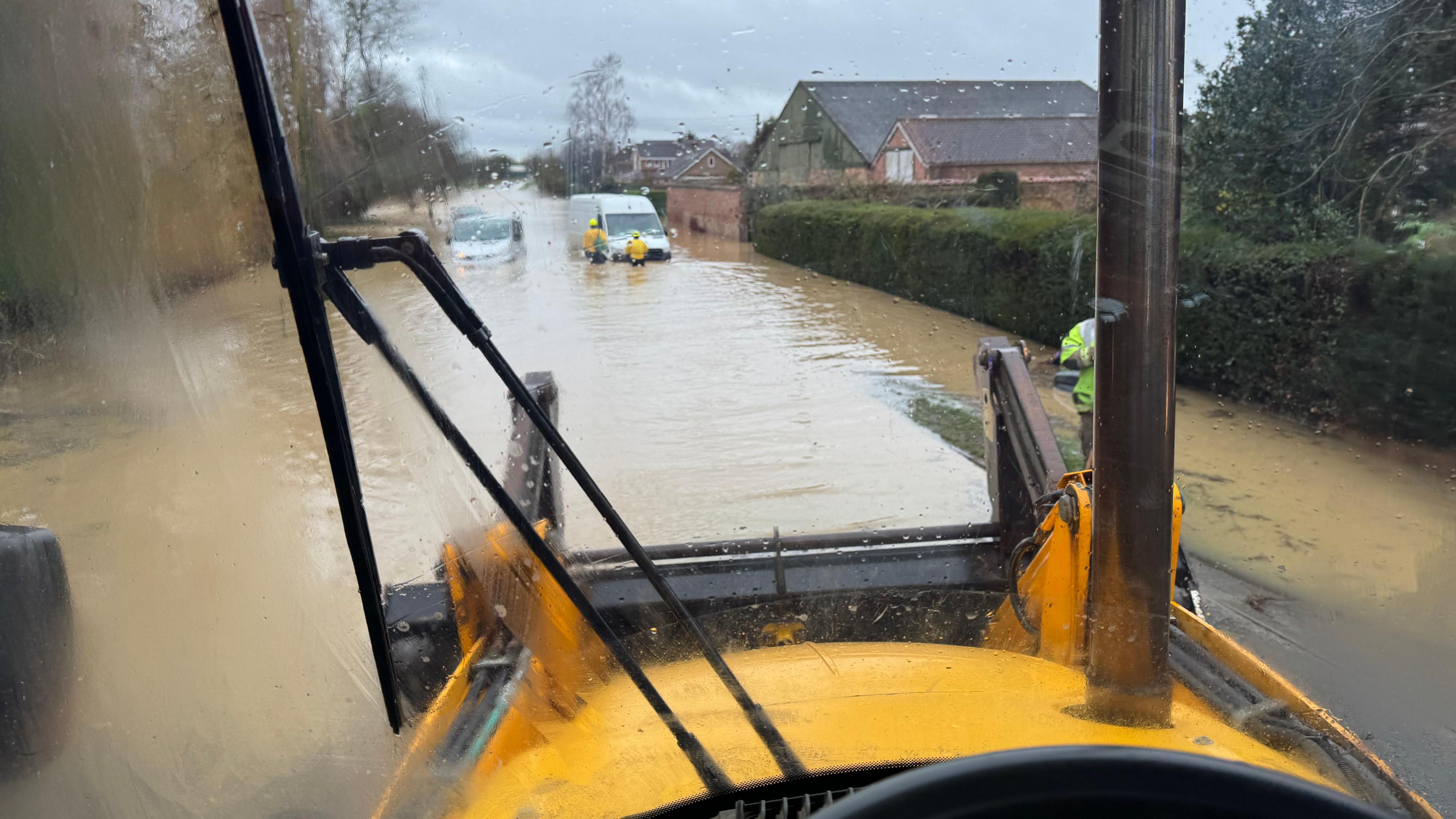 A photo from inside a yellow digger. It is driving through deep brown floodwater. There are two vans in the distance, with water up to their windows. 