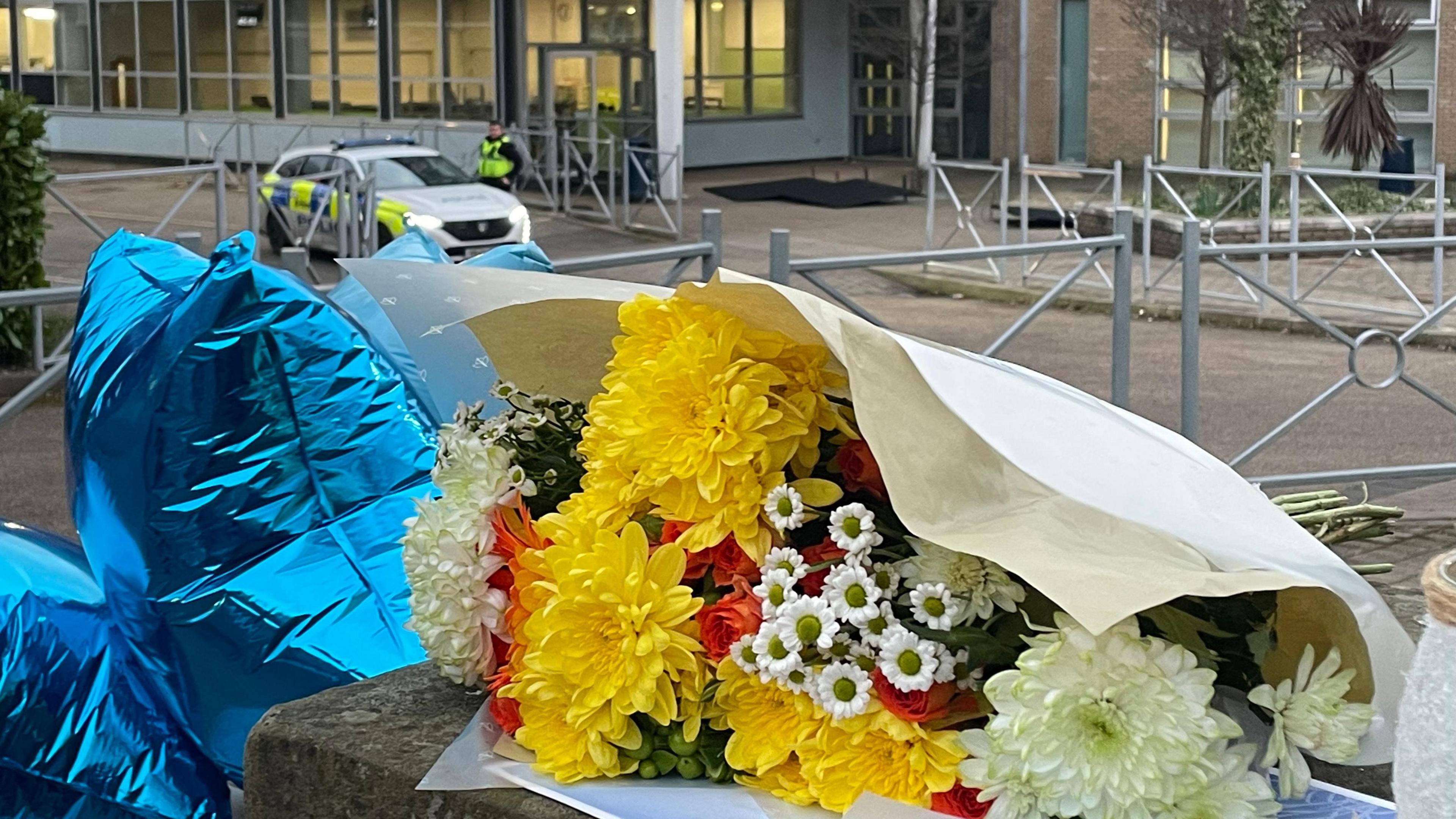 Flowers and balloons left on the wall outside the school building. A police car and officer can be seen in the background, near the entrance.