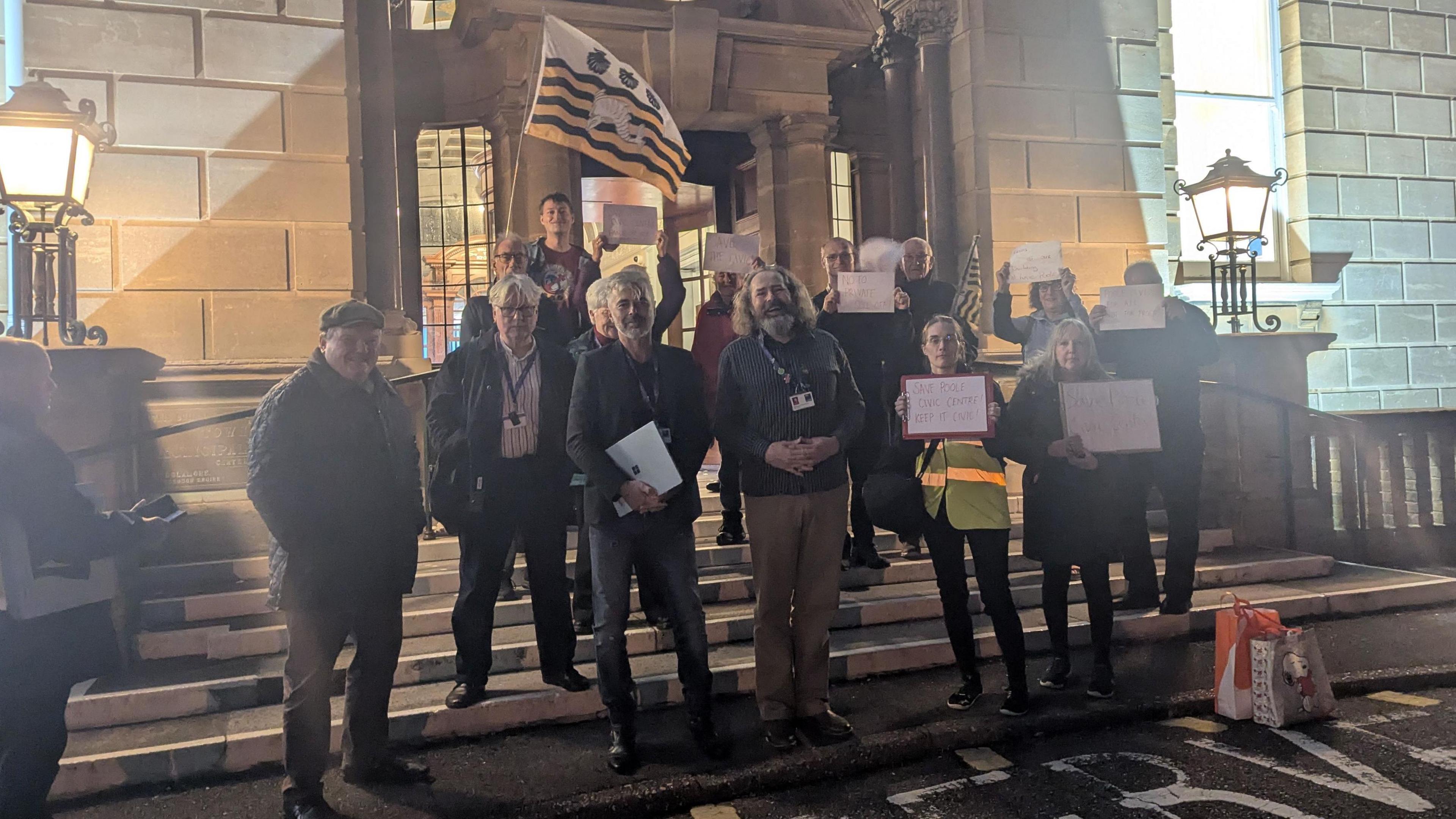 Thirteen men and women standing on the steps of Bournemouth Town Hall - some are holding up sheets of A4 paper but it is not possible to read what's written on them. It is after dark and the scene is lit by light shining from inside the building and large ornate lamps mounted on pillars either side of the steps