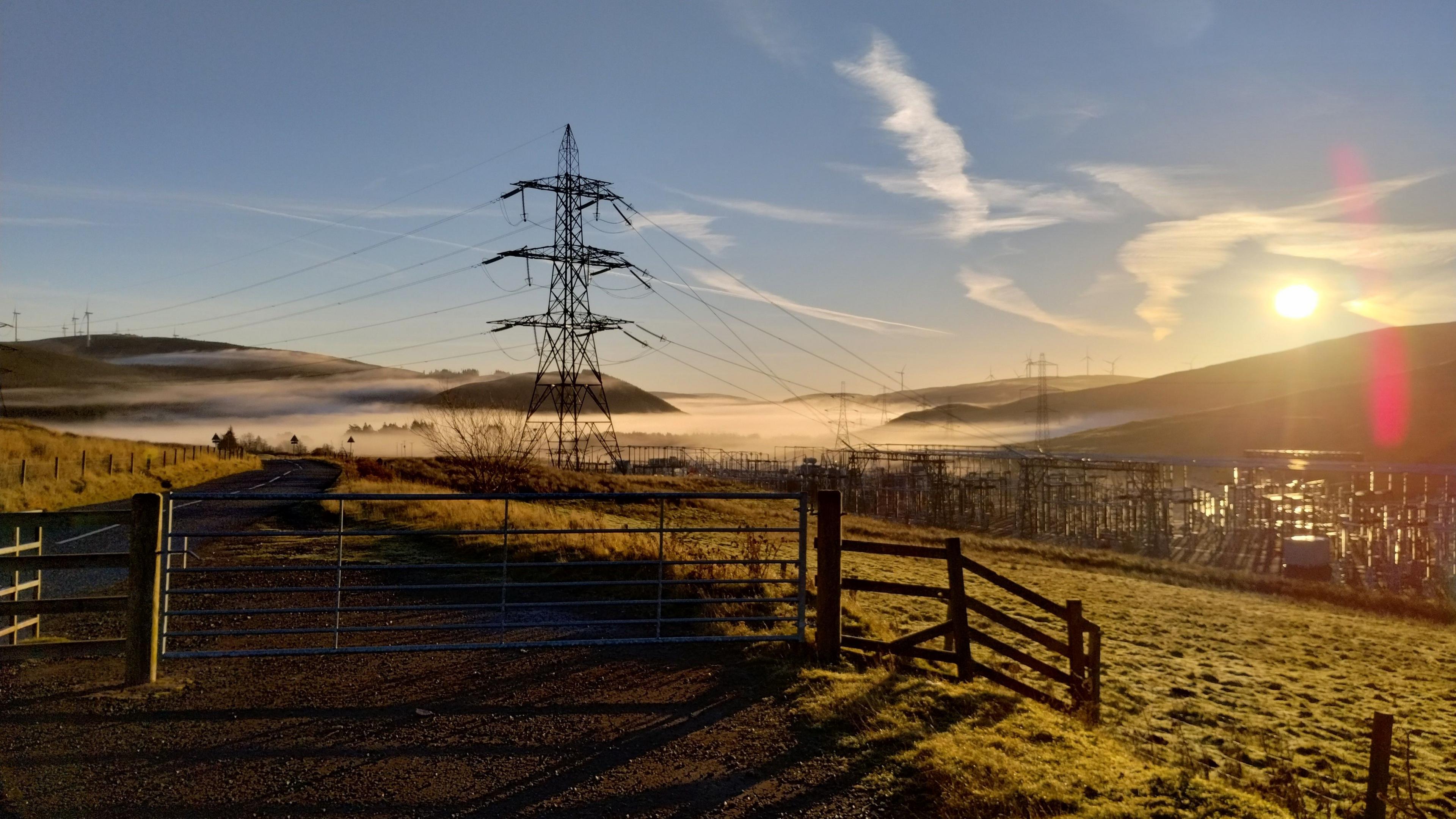 Some late autumn sunshine on a south of Scotland hillside with an electricity pylon in the foreground and wind turbines in the distance