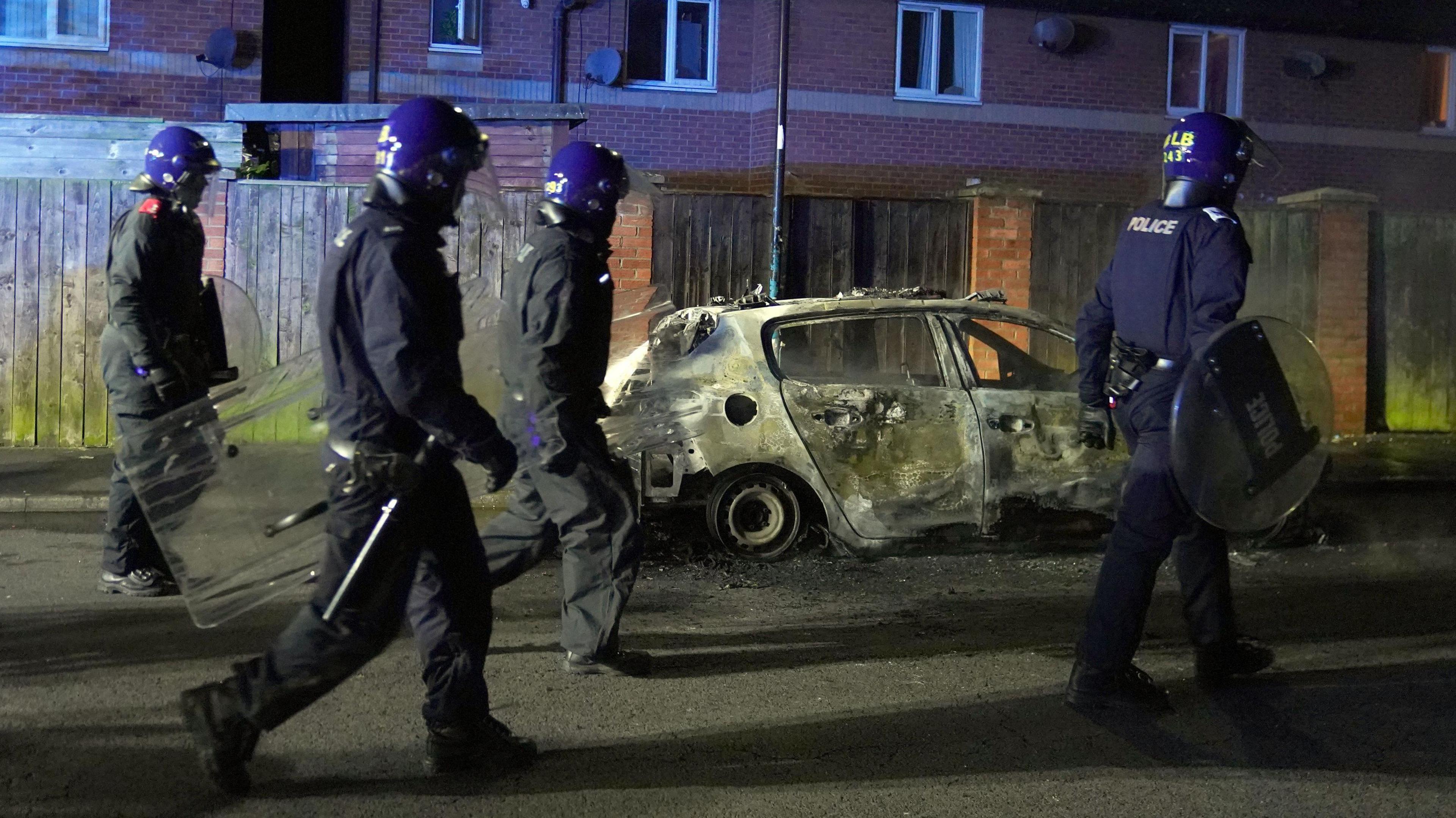 Four police officers with riot shields walk past a burnt-out police car