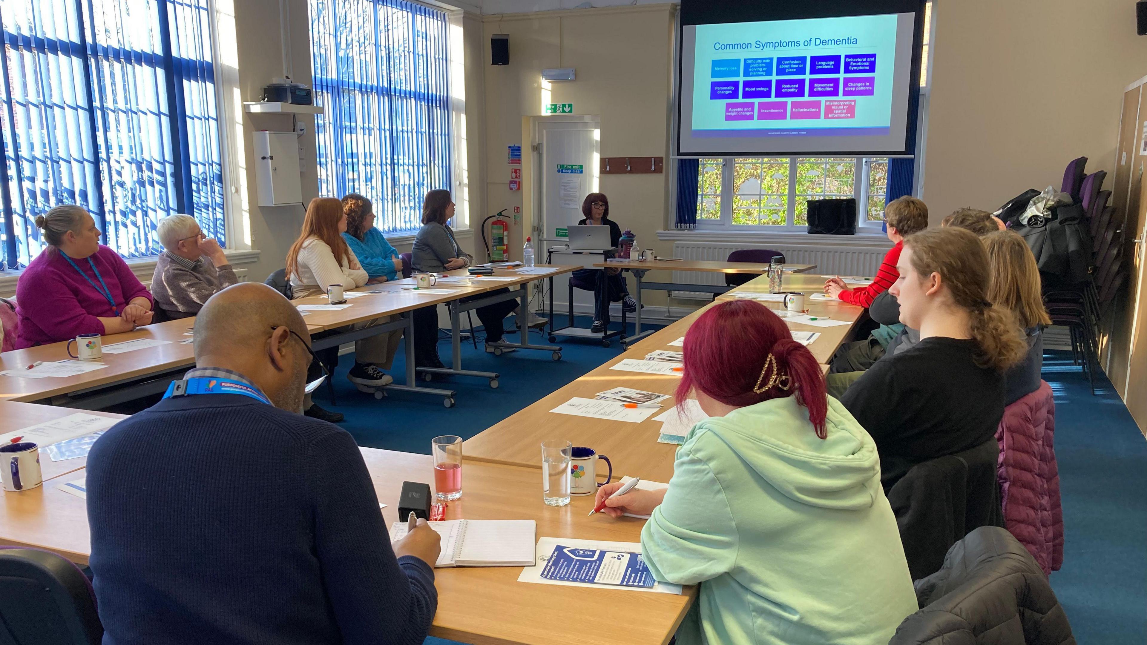 A number of people are sitting at tables placed together to form a rectangle. A projector screen at the end of the room is showing a slide titled: "Common Symptoms of Dementia". Some of the people are taking notes.