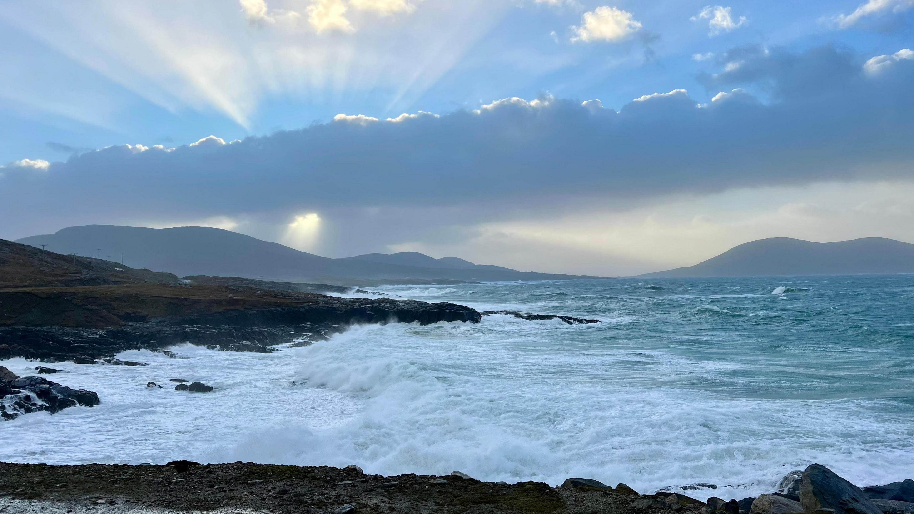 Waves crash against the shore on the Isle of Harris. The sun is hidden behind dark clouds in the shot.