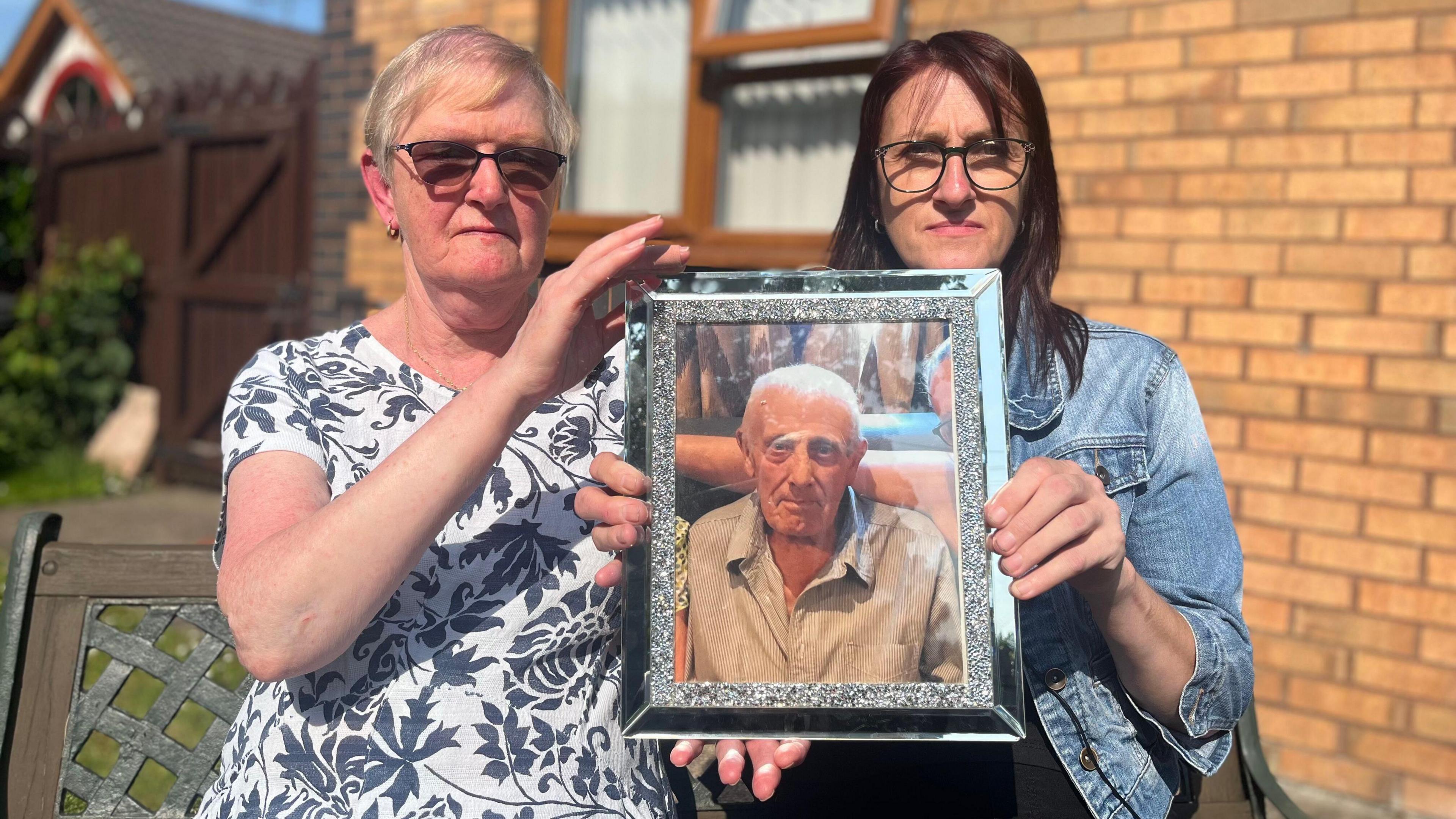 Two women hold up a silver framed picture of an older man.
The woman on the left, Susan Holmes, is wearing a black and white floral patterned top and dark glasses and has short blonde hair while the woman on the left, Lisa Jones, has long dark hair and is wearing a denim jacket and glasses. The man has short white hair and is looking directly at the camera while wearing a striped shirt. Behind them you can see the wall and window of a house.