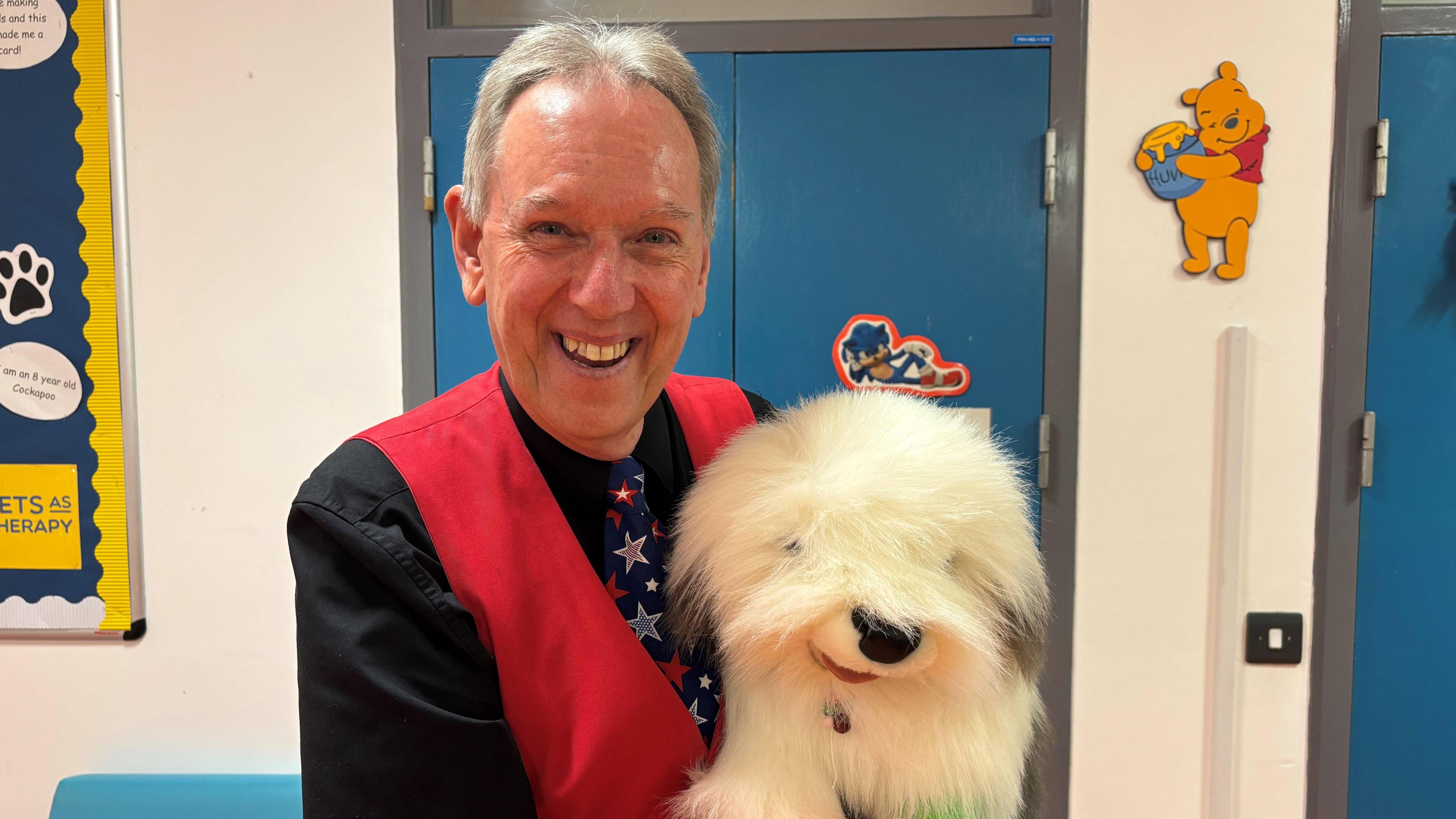 David Oakley the magician has short grey hair. He is wearing a black shirt and red waistcoat. His tie his navy with stars on it. He is holding a puppet of a white old English sheep dog