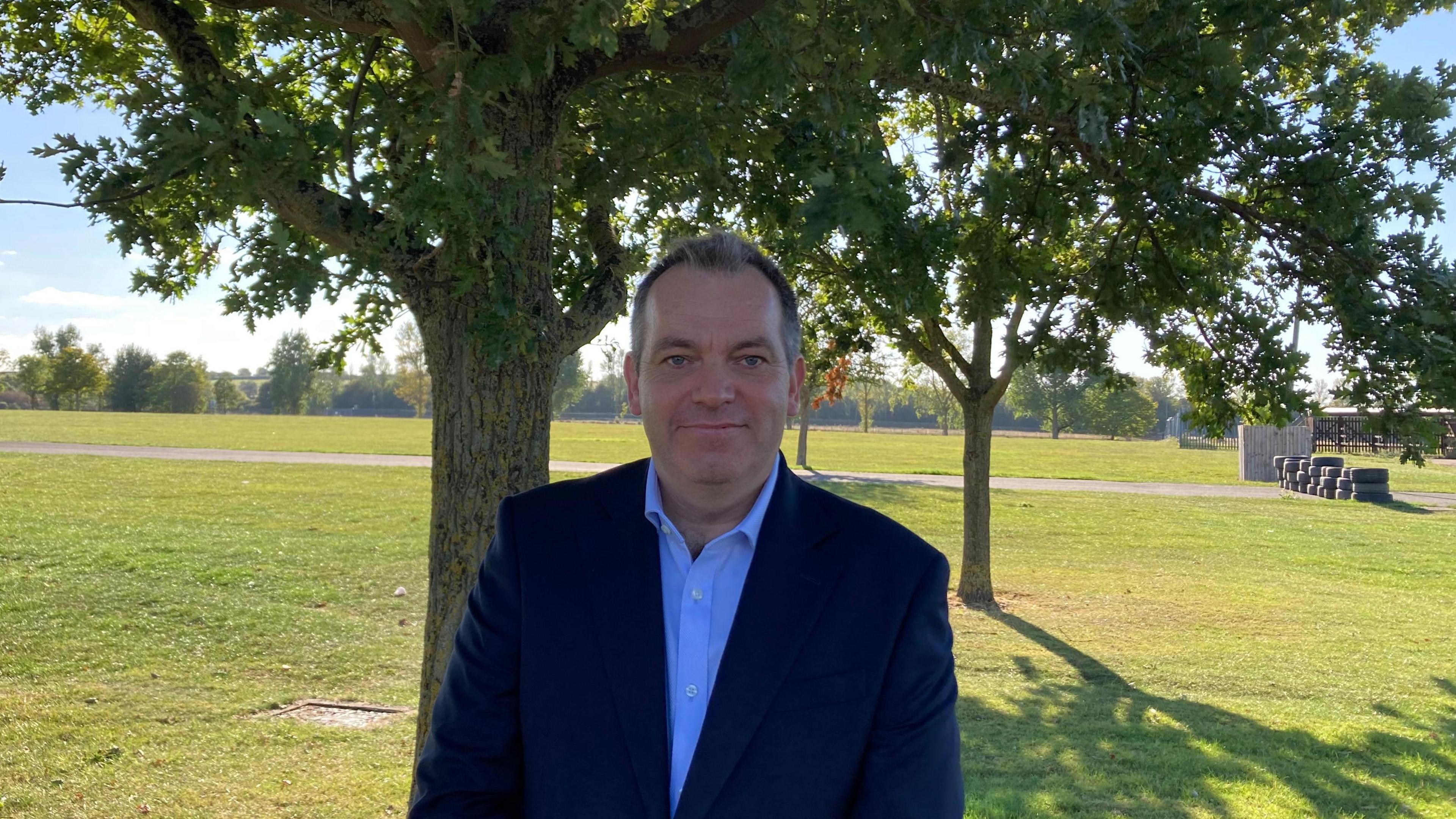 Ashley Butterfield is standing in front of a tree in a grassy area, wearing a blue short and dark blue suit