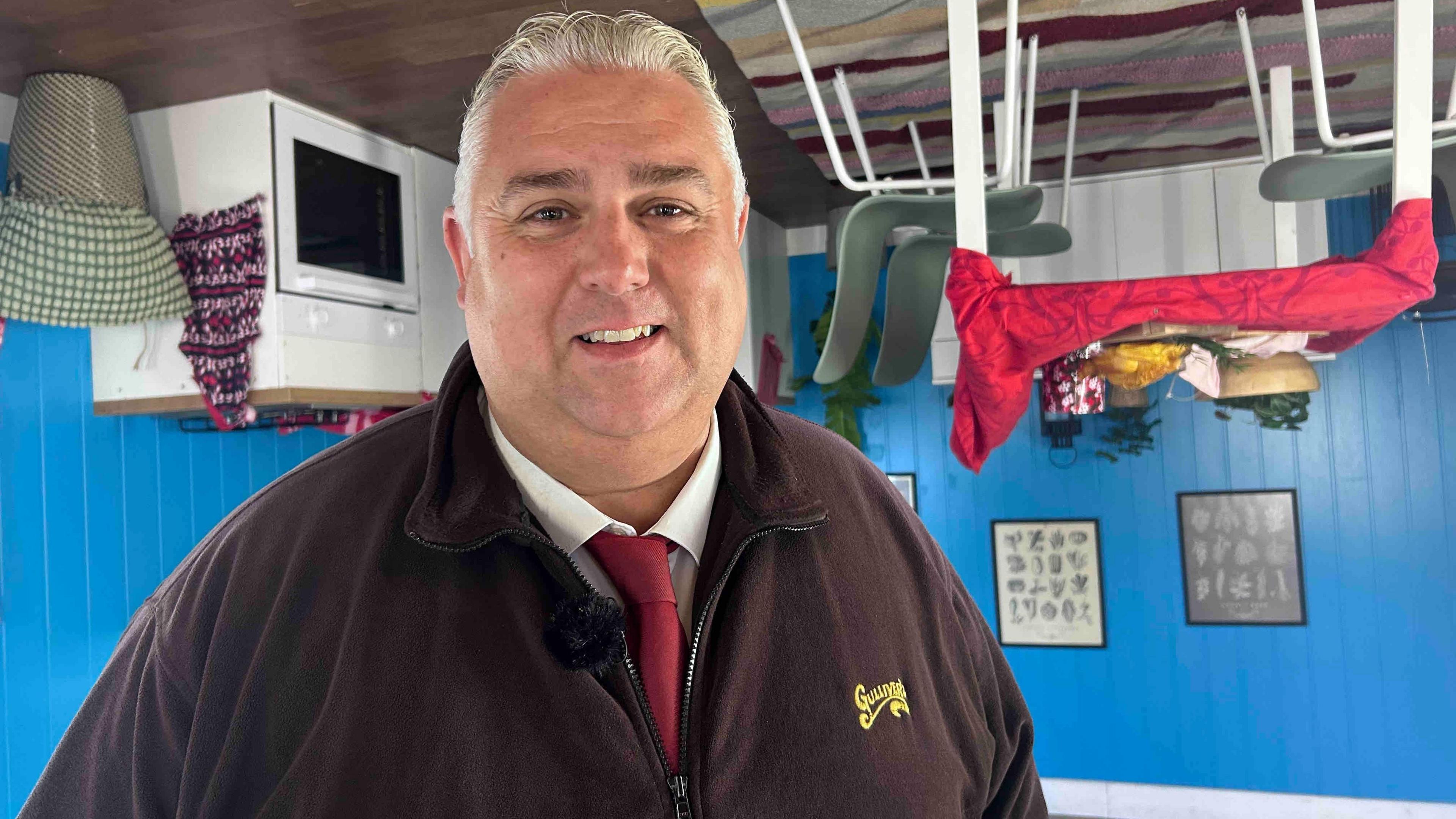 A man wearing a white shirt and red tie has a black fleece with the words "Gulliver's" written in yellow stitching.