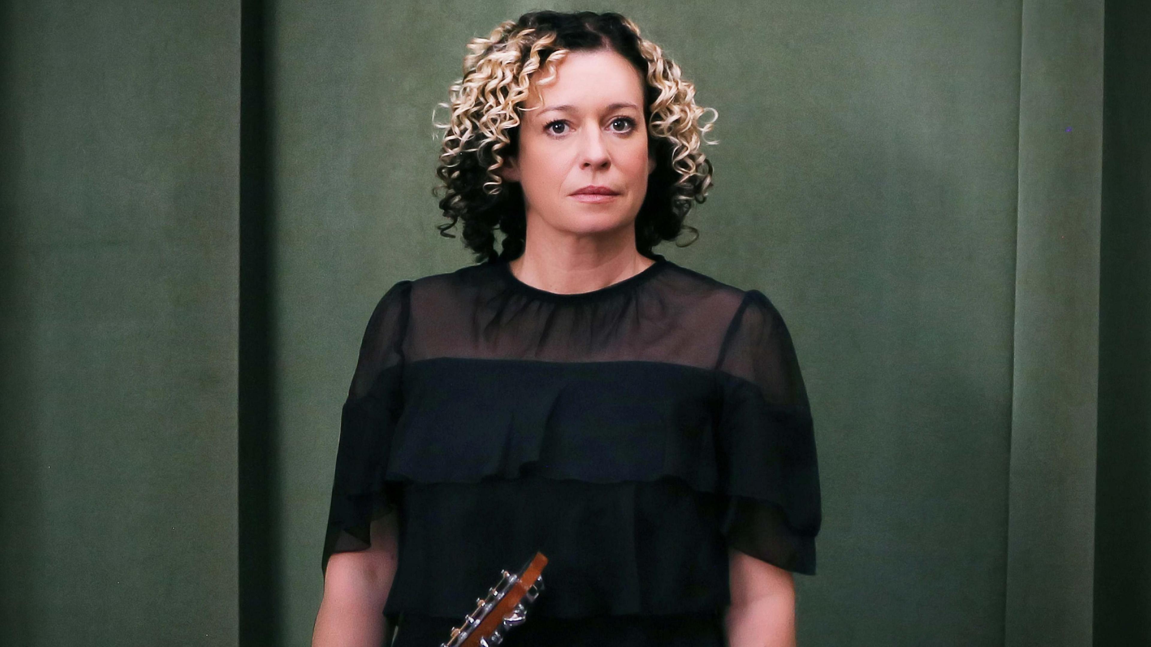 A woman with curly hair and a black top holds a guitar while looking into the camera.