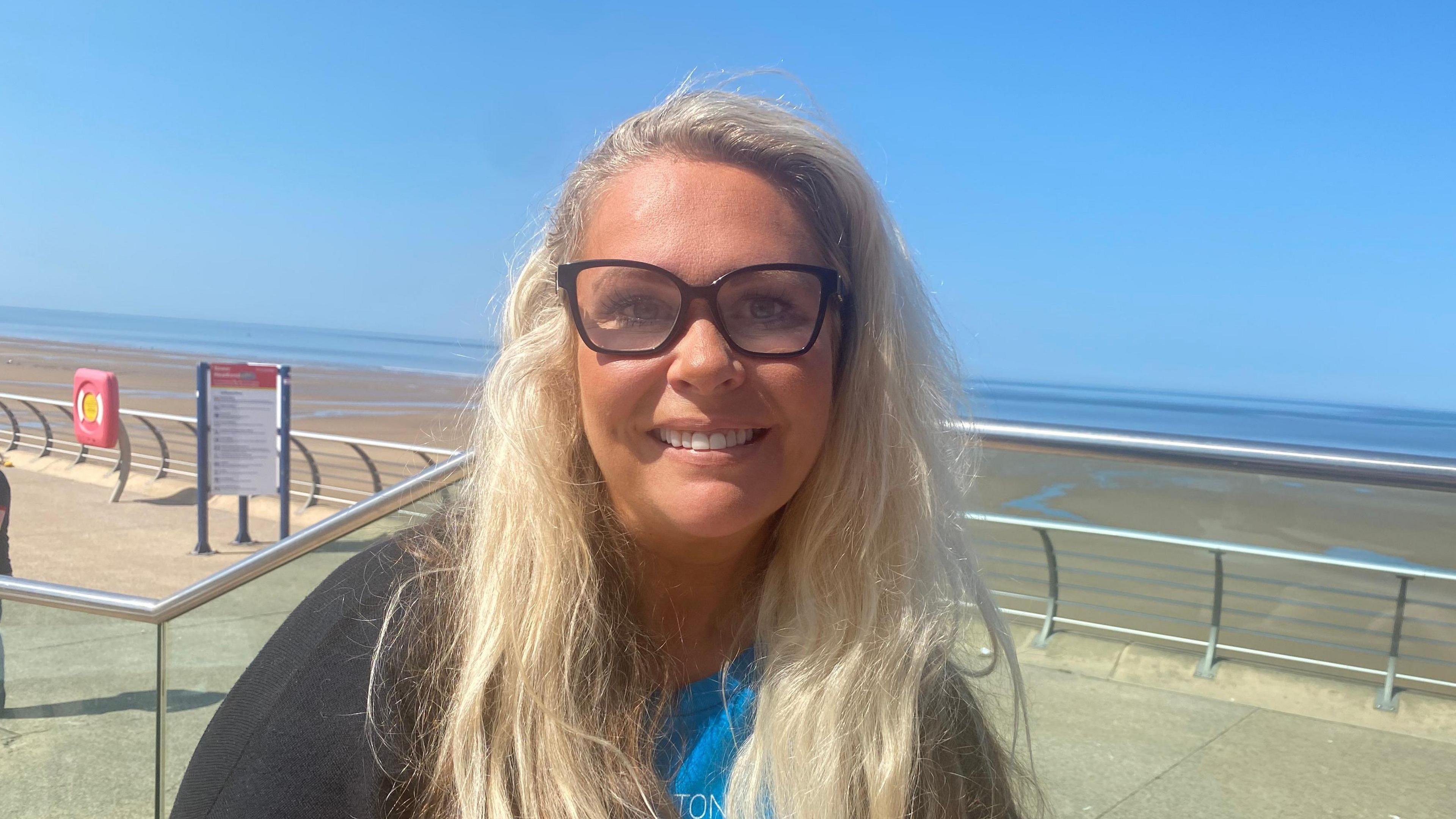 A closeup photo of a woman with blonde hair and black rimmed glasses, smiling to the camera in front of Blackpool Prominade