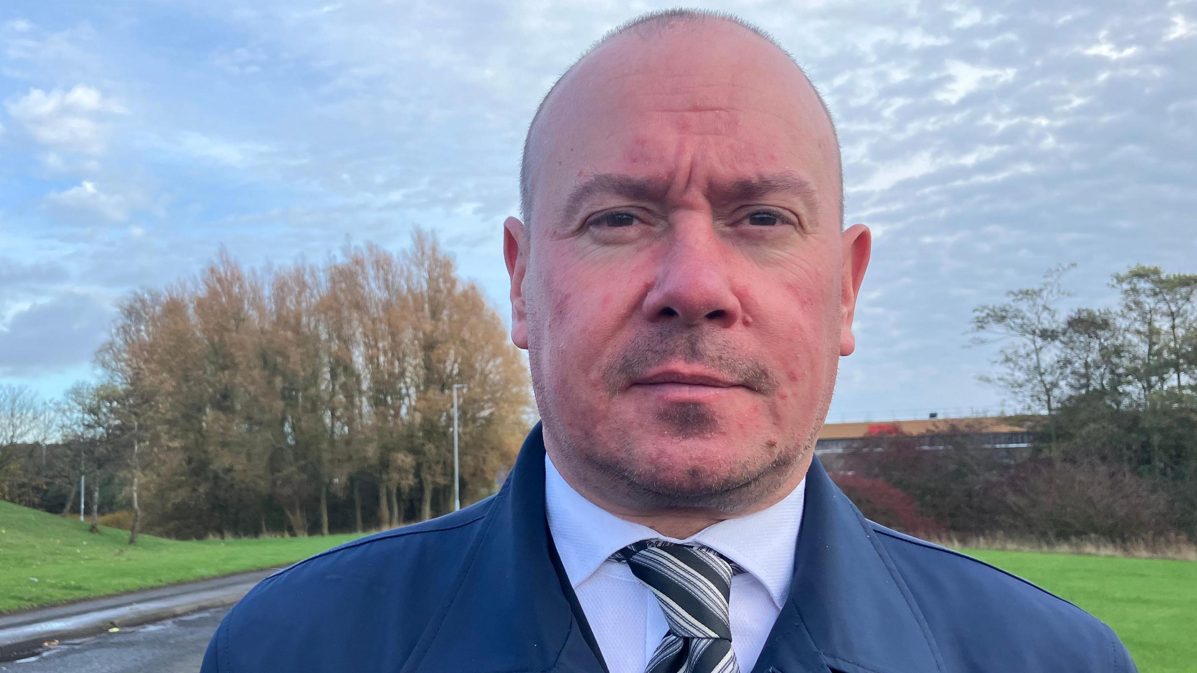 Steve Bush. He is wearing a tie and a smart blue coat. He has a five o'clock shadow and is bald.