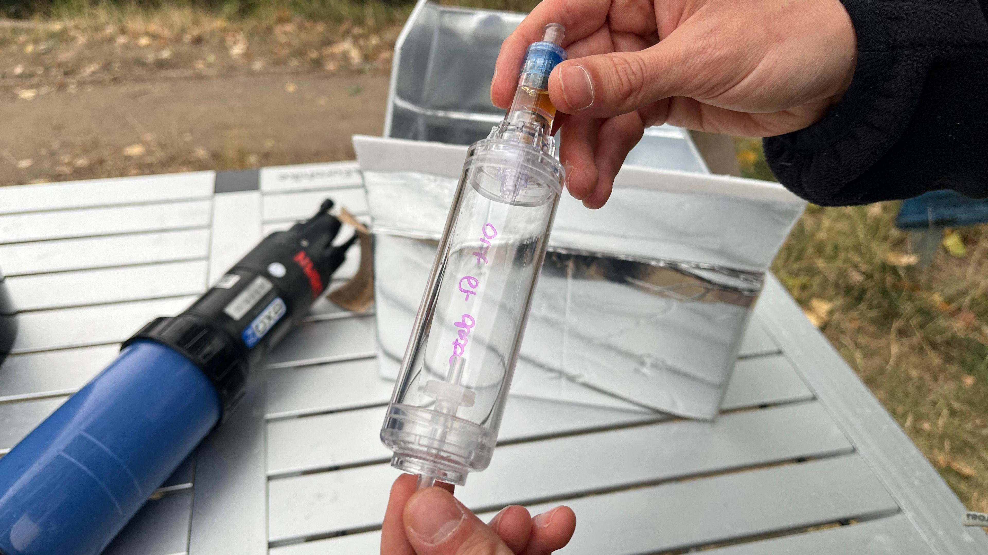 A person's hands holding a glass cylinder that is narrower at the top end. It's being held over a metal trestle table, and under the table you can see grass and dirt. The person holding the cylinder is wearing a black fleece.
