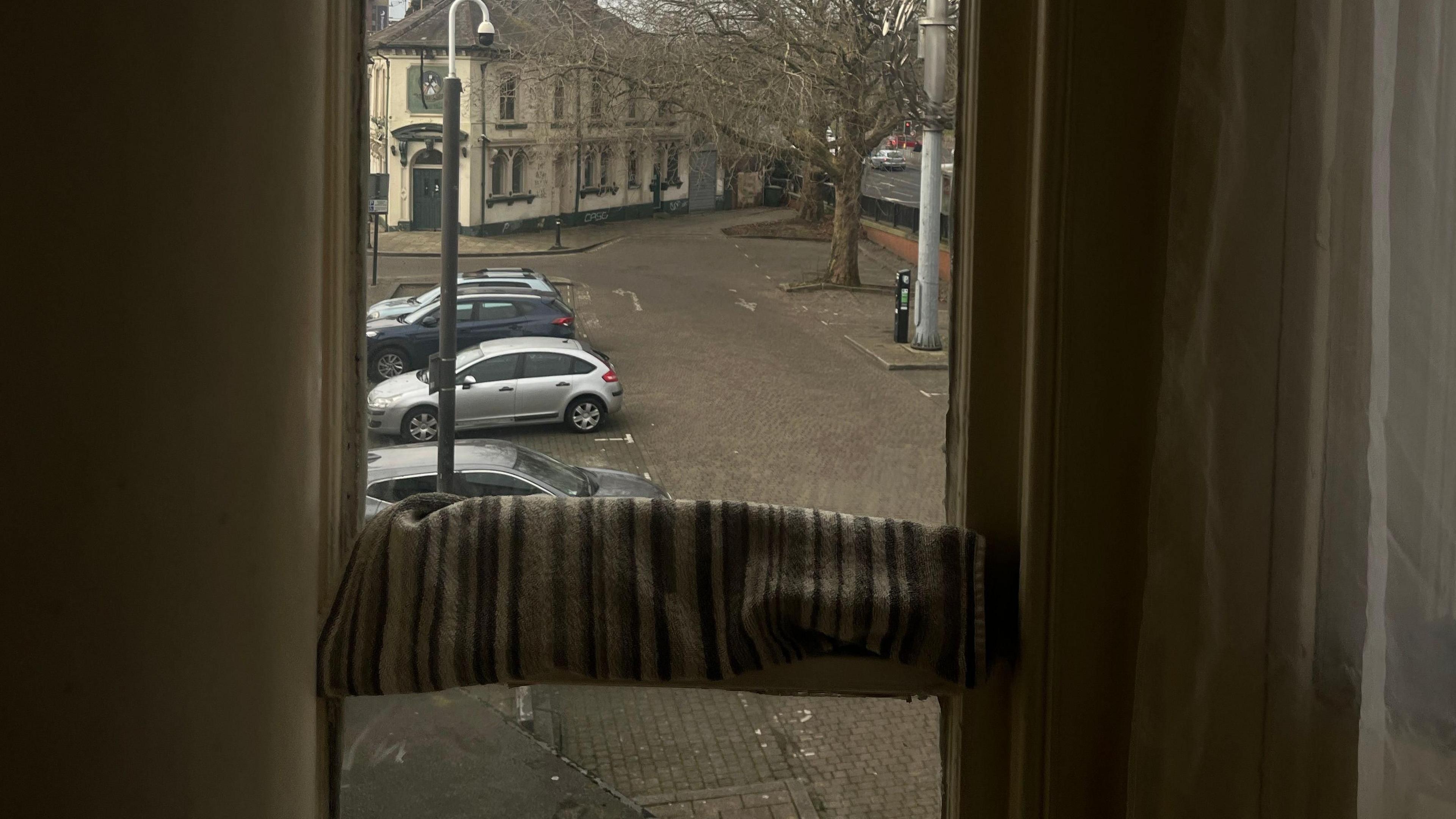 A narrow window with the view to a car park. There is a cream building and trees with no leaves. There are four cars parked. The window has a towel across the middle that is being used to block draughts. 