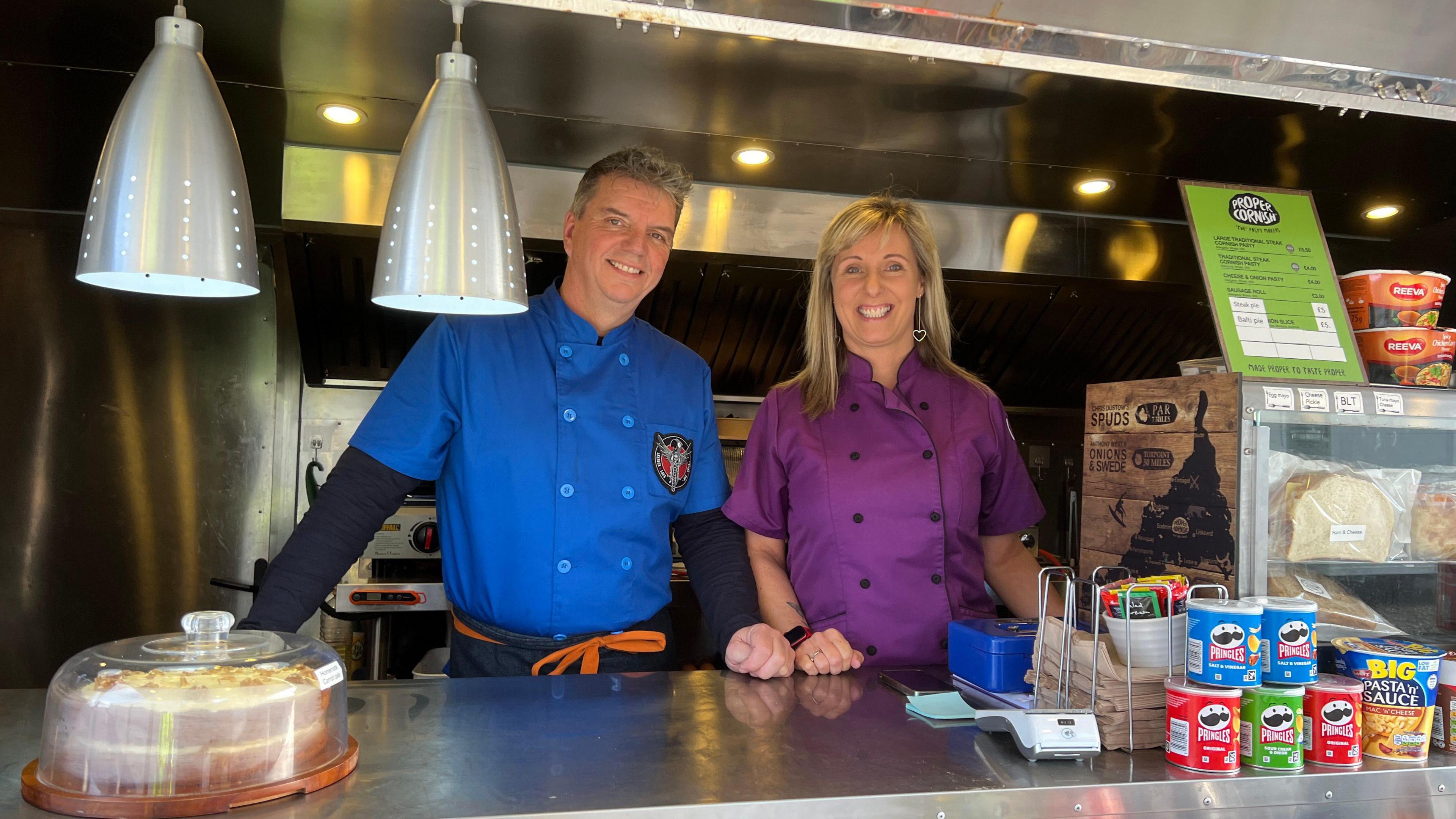 A man in a blue chef uniform stood smiling in a food van with a big cake and around it. A women in a purple chef uniform is stood next to him. There's pasties and toasties and Pringle tubes in the counter.