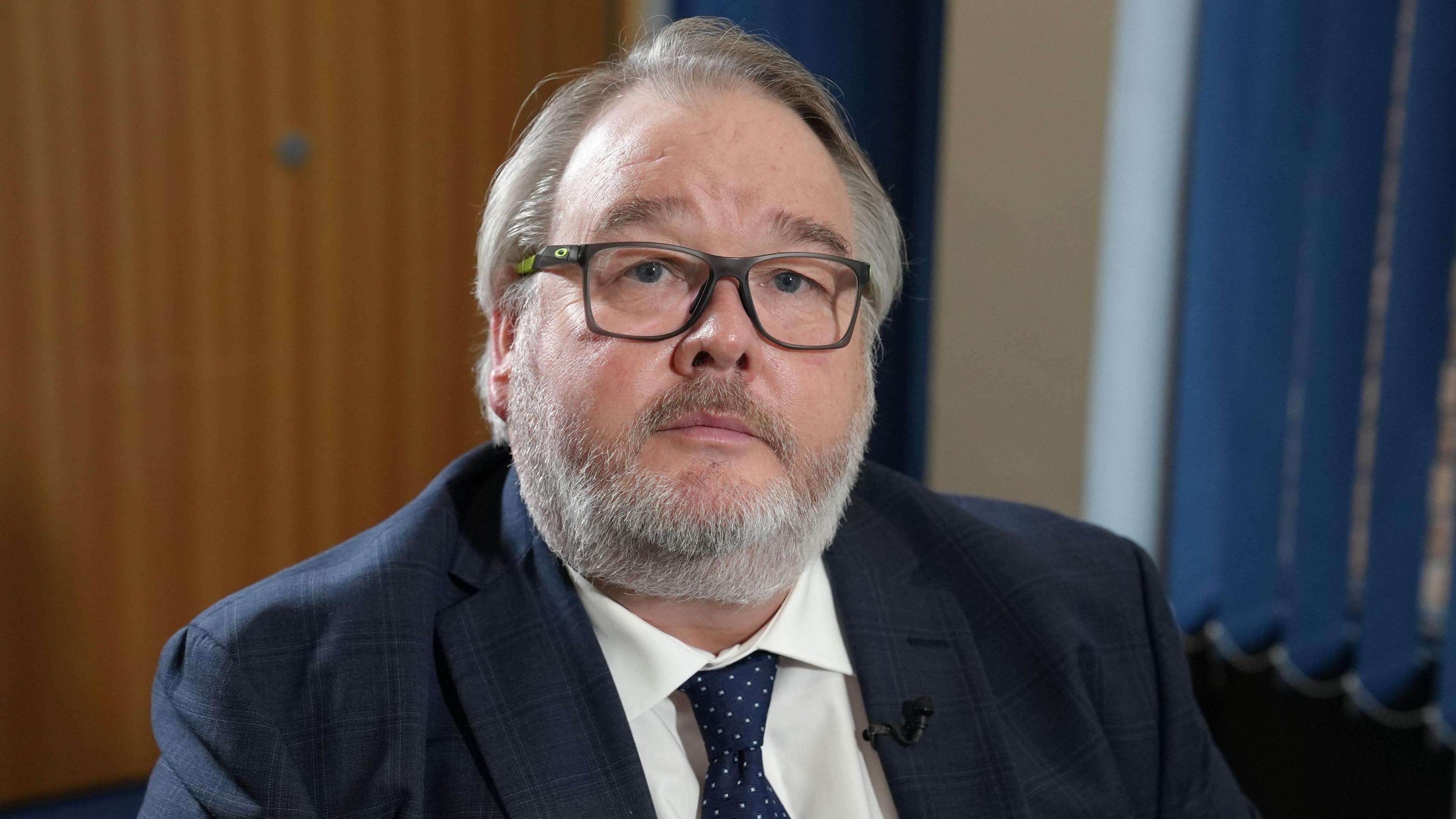 Christopher Mills mewn crys wen, siaced ddu a thei smotiog. Yn gwisgo sbectol a ddim yn gwenu.