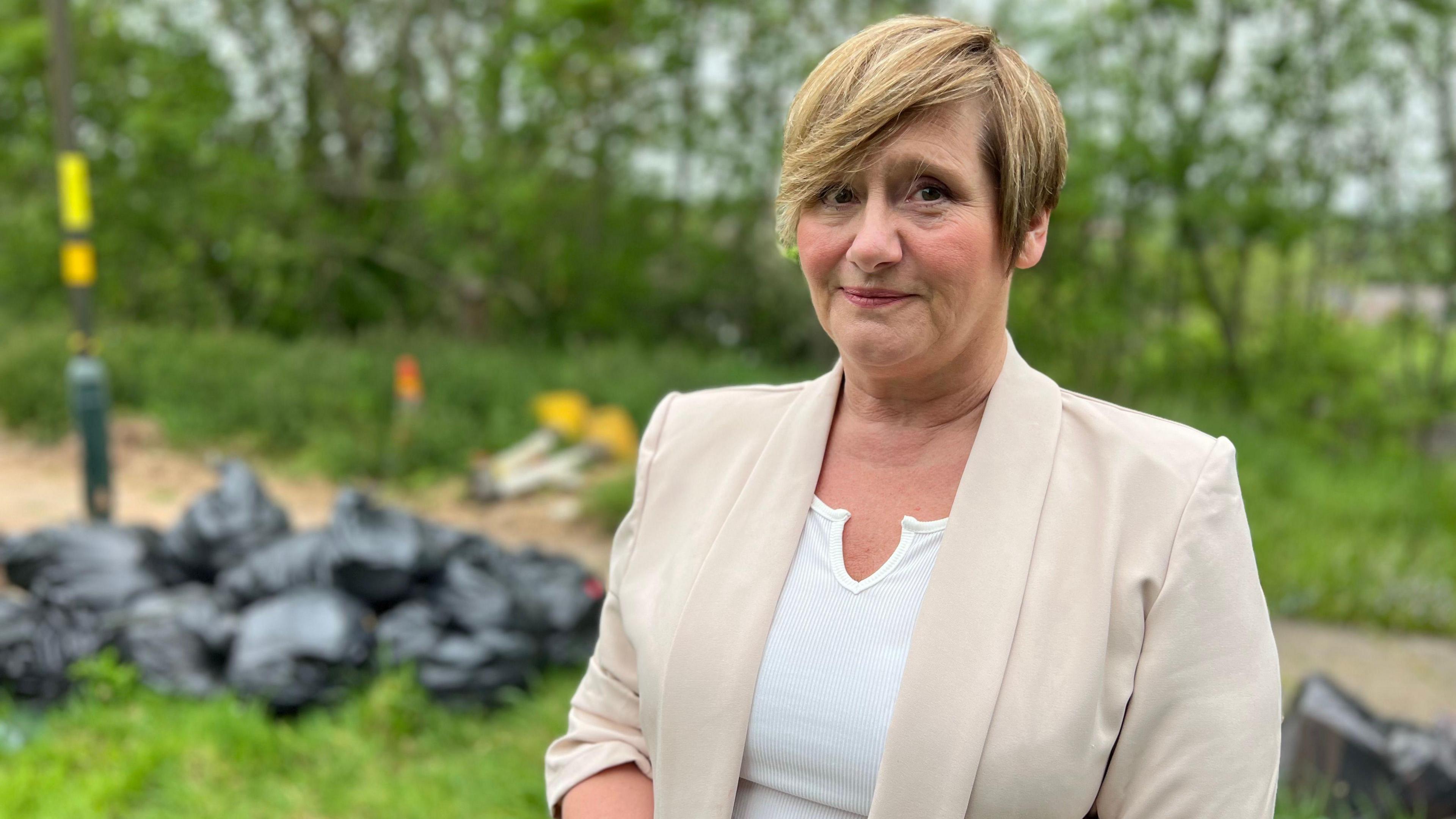 Candy Manders has short honey blonde hair and is wearing a beige blazer and white top. She is standing in front of green grass which has a pile of black binbags on it.