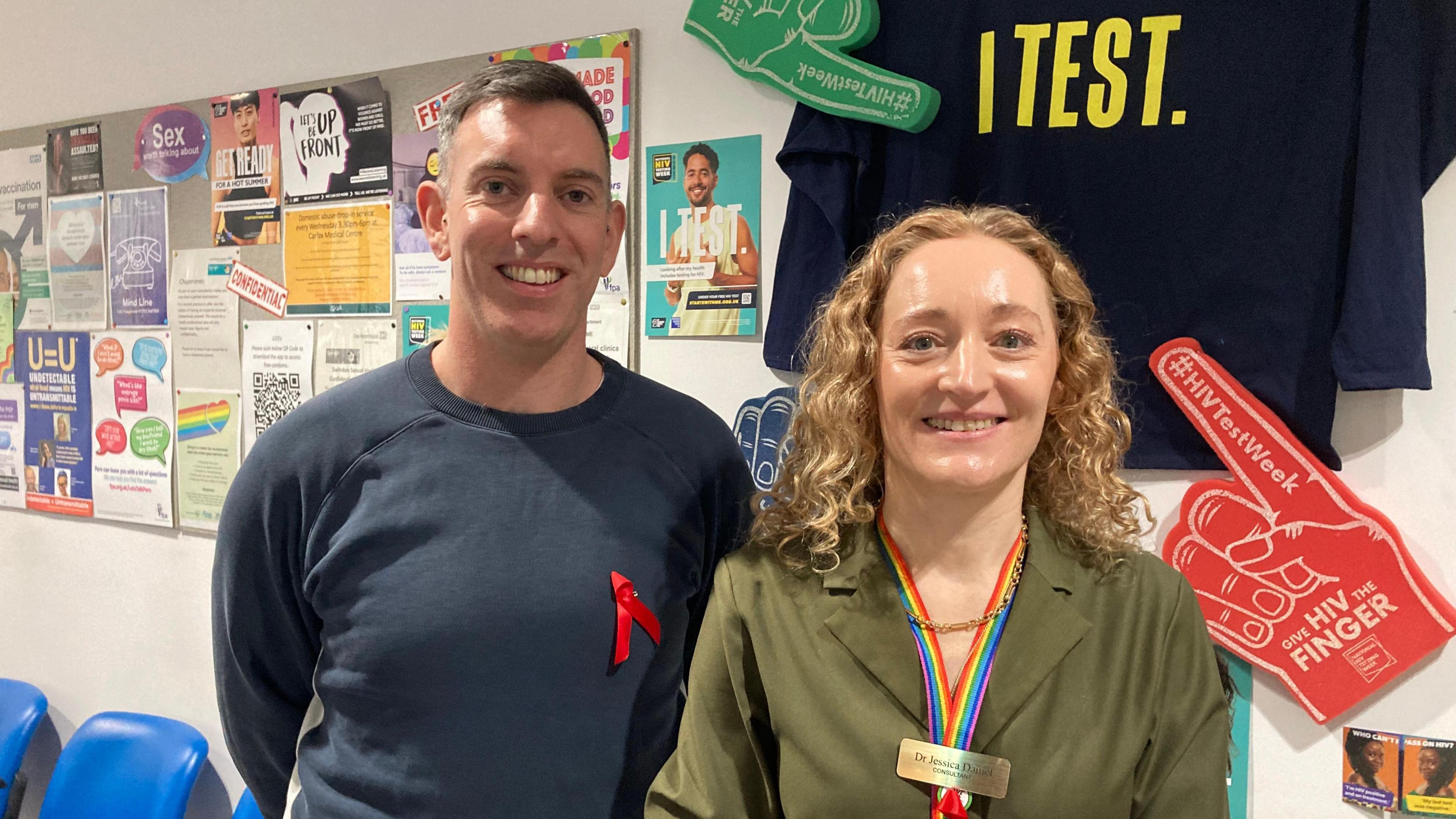 A man and a woman standing next to each other smiling at the camera. On the wall behind them is a foam finger, with the words "HIV Finger" written on it, a navy T-shirt with the words "I Test" and a bulletin board with various notices.