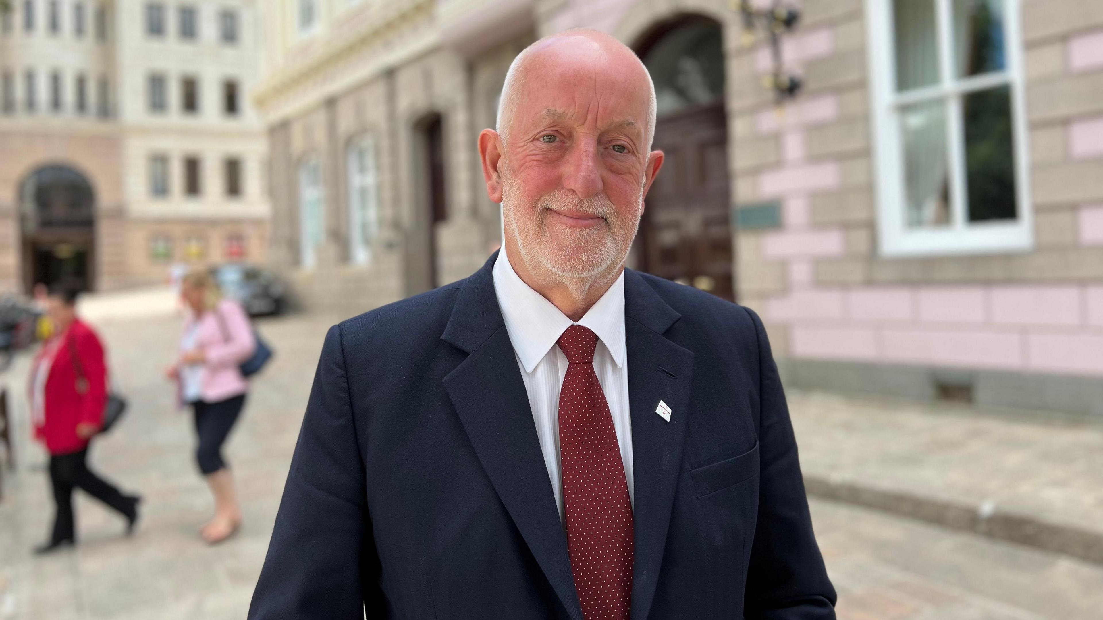 A man wearing a suit and tie standing in front of a government building. Background is blurred.