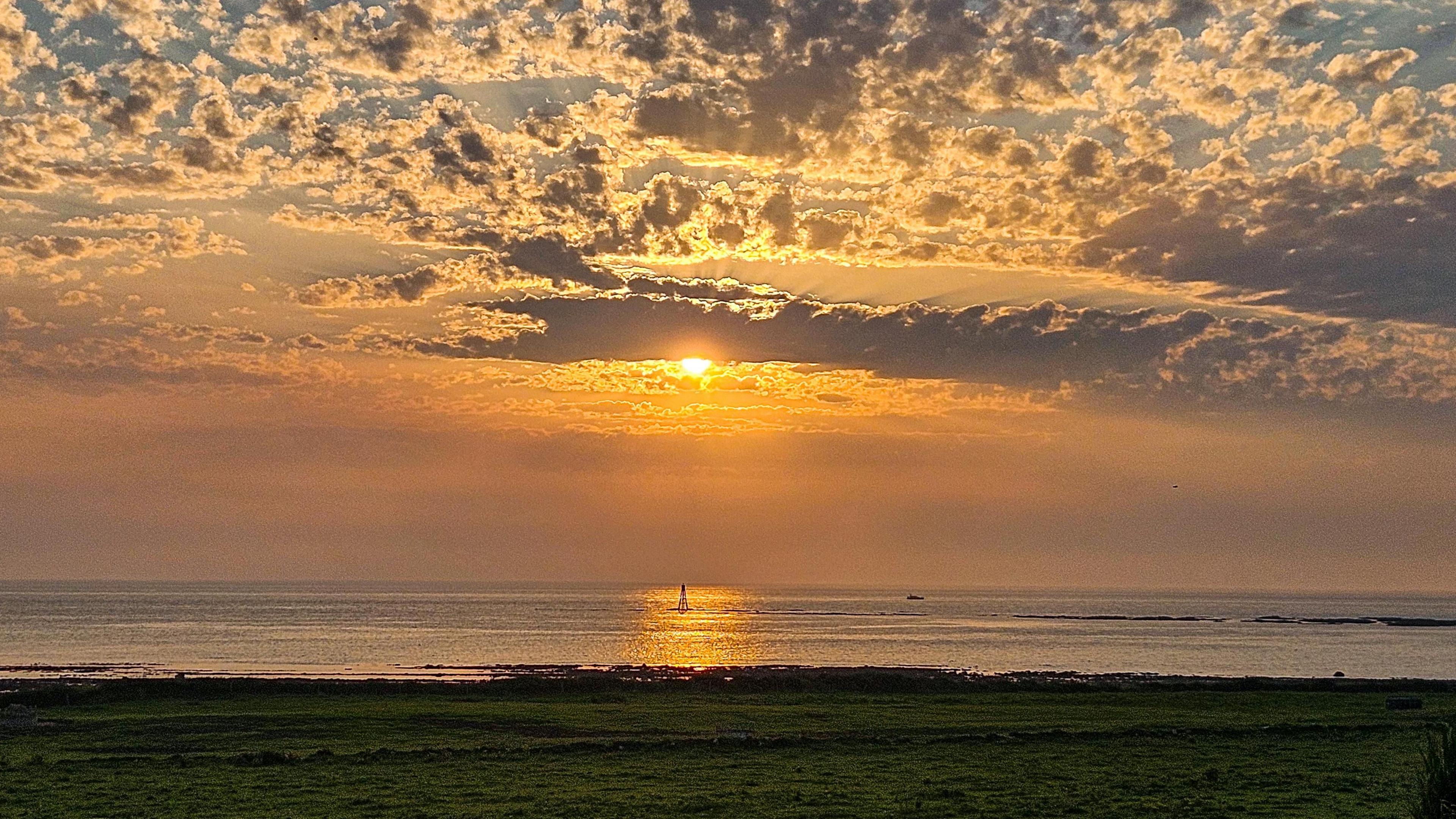 Landscape image of the sun setting over calm waters at Turnberry beach. The sky is an orange, yellow and blue ombre with the sun peaking from behind clouds.