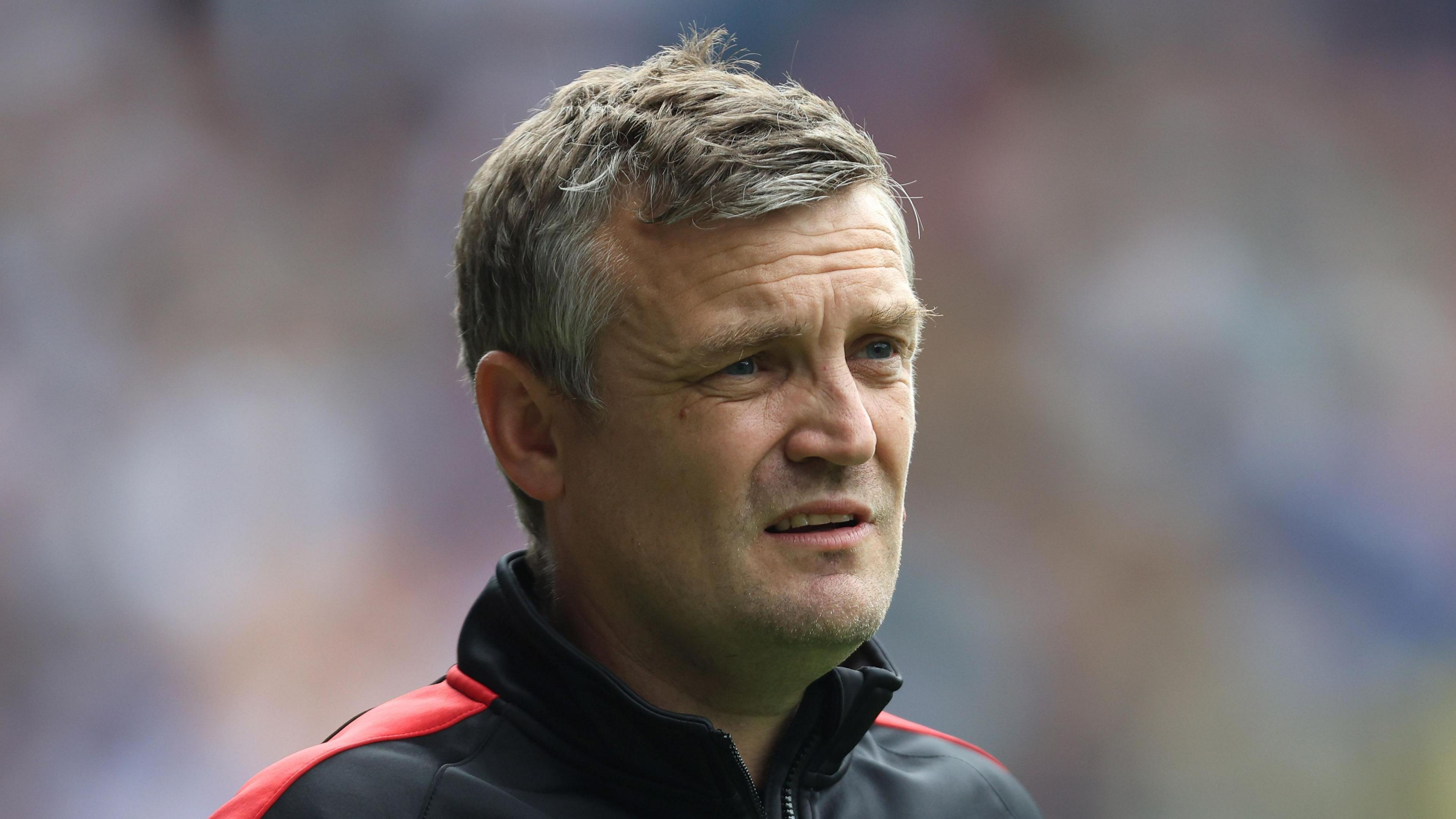 Lincoln City head coach Michael Skubala wearing a tracksuit top looks on with his eyes narrowed.