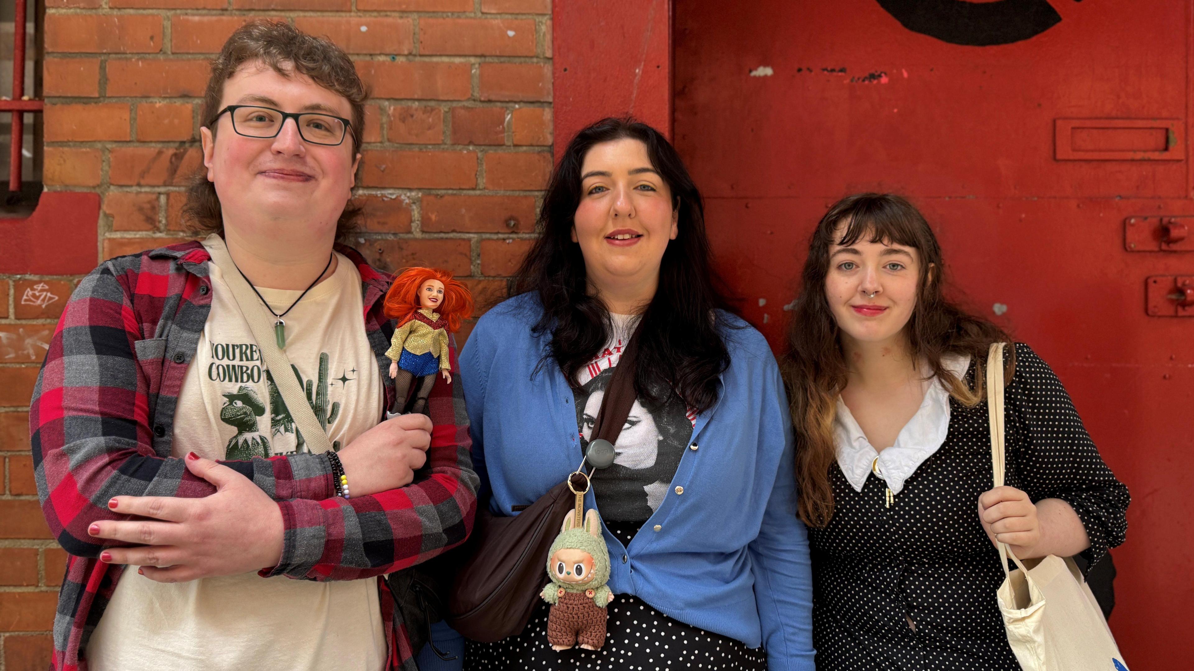 A man and two woman stand against a wall and a red door. The man (left) wears a red checked shirt and a white top. He has curly brown hair and glasses. The woman (middle) has a blue cardigan and long black hair. The final woman (right) wears a black spotty dress. She has long brown hair.