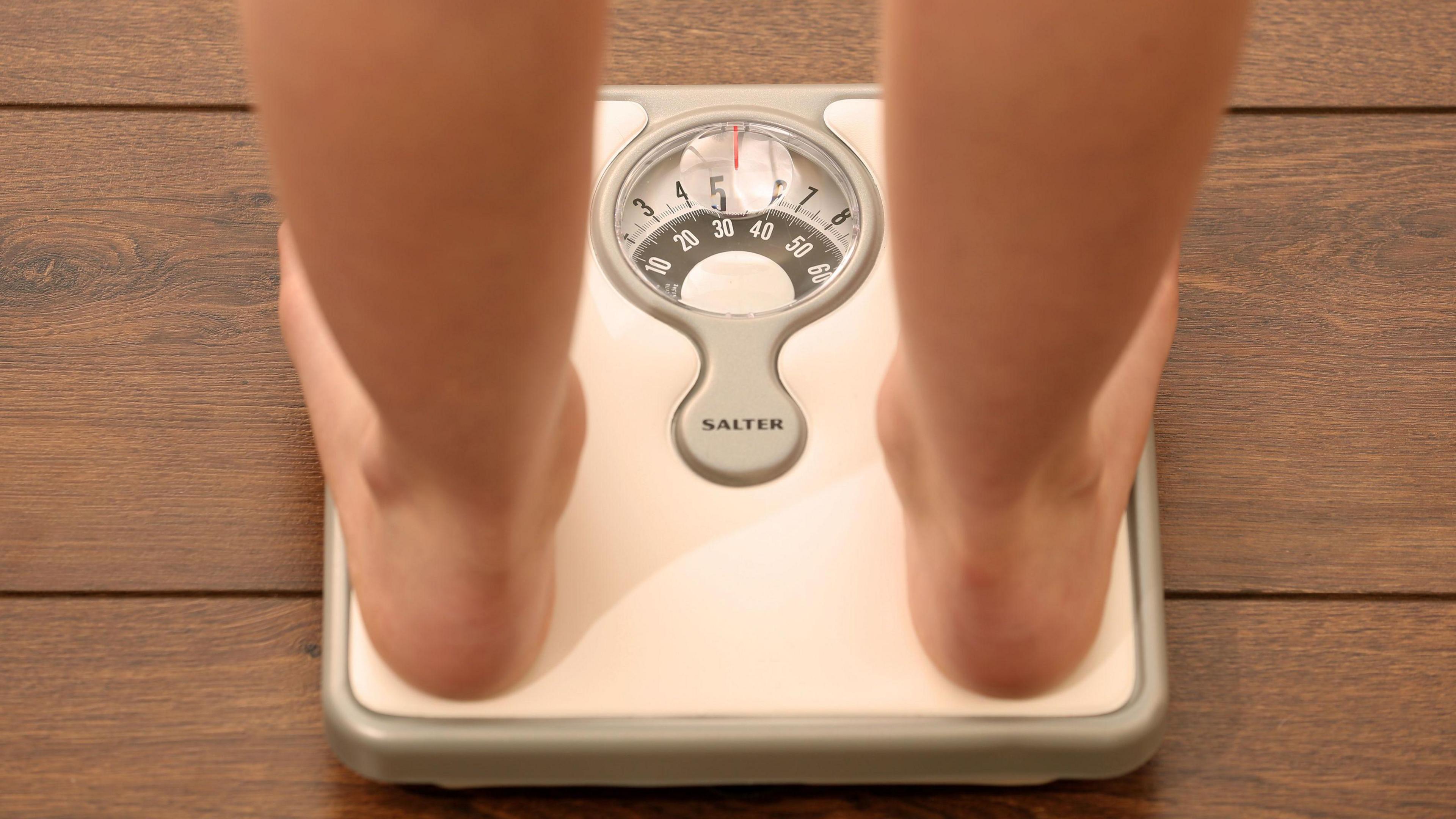 A pair of bare feet on white scales. The scales are on a brown wooden floor.