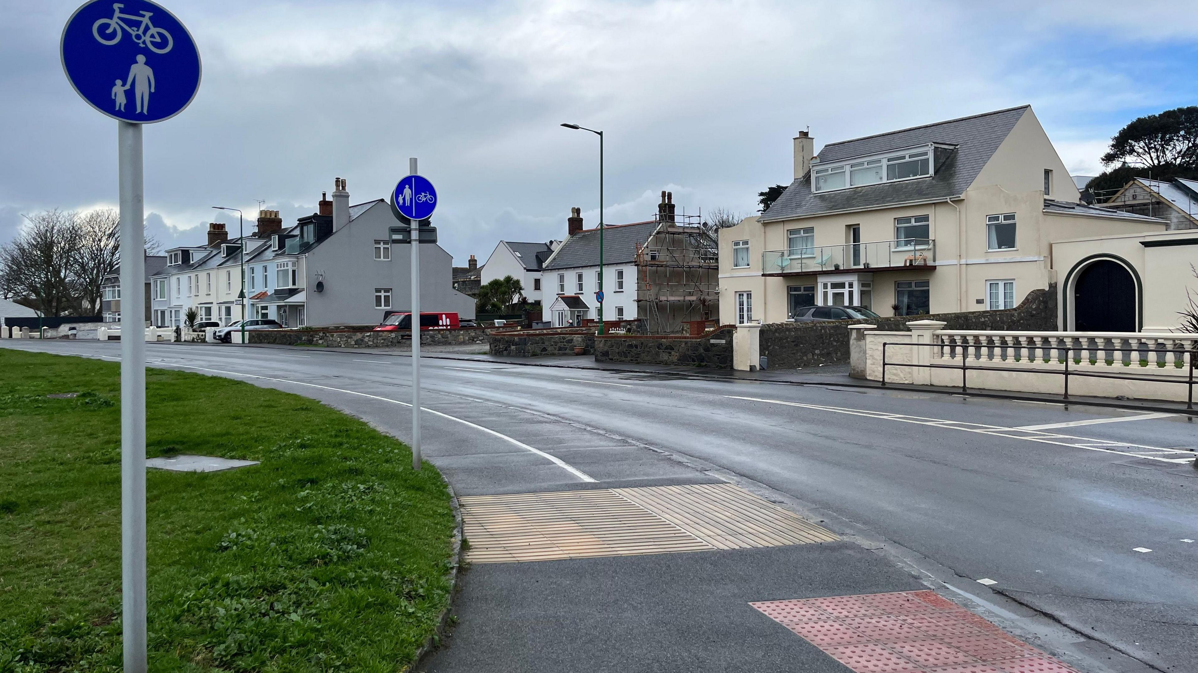 A sign with a pedestrian and child, with a bike above. In front of a grassy verge and a pavement with some blister paving and a cycle path. 