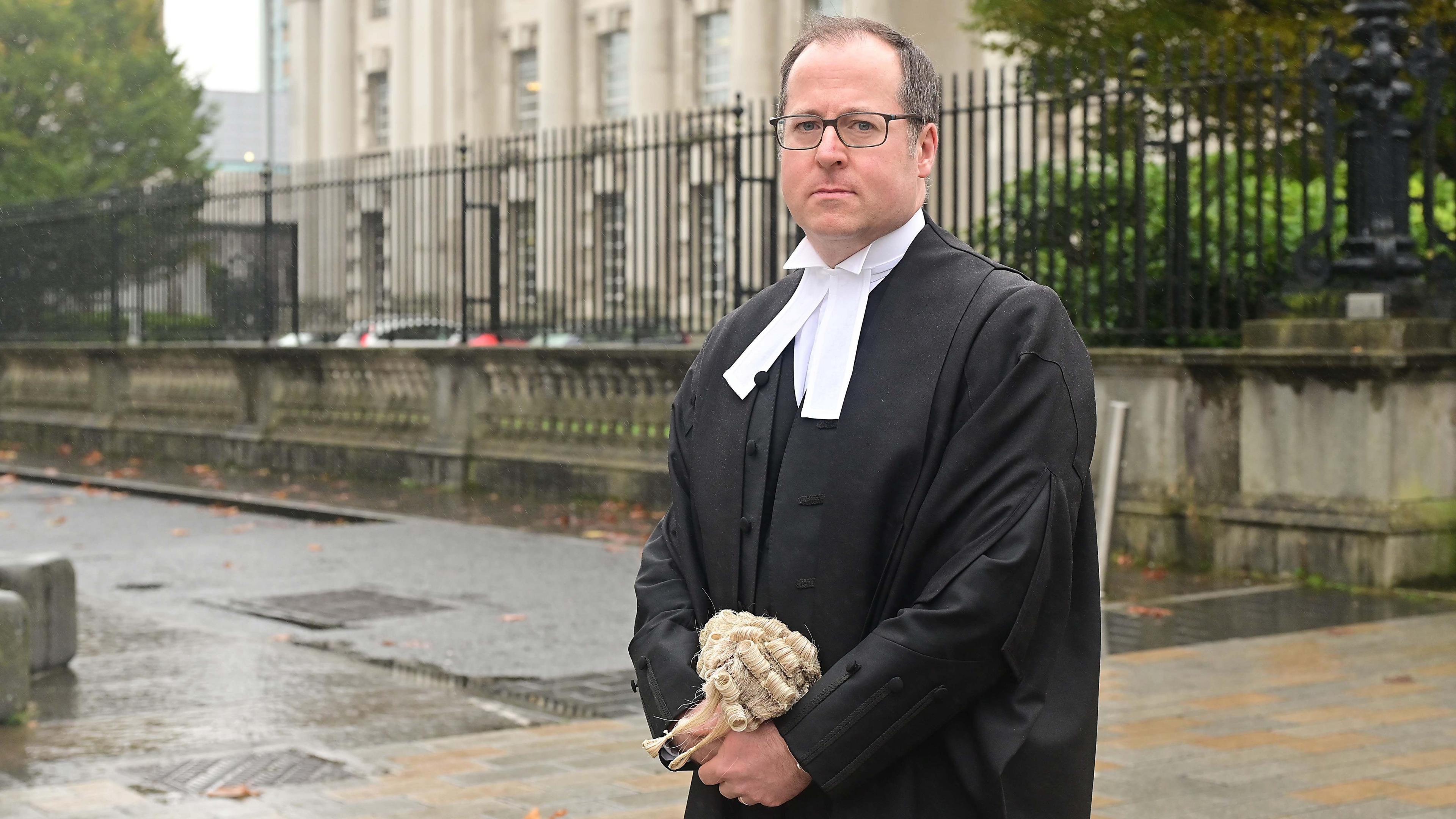 Donal Lunny KC has short brown hair and is wearing square dark framed glasses. He is wearing black robes and a white shirt underneath. He is holding his barrister wig in his hands and standing in front of the black railings at the courthouse. 