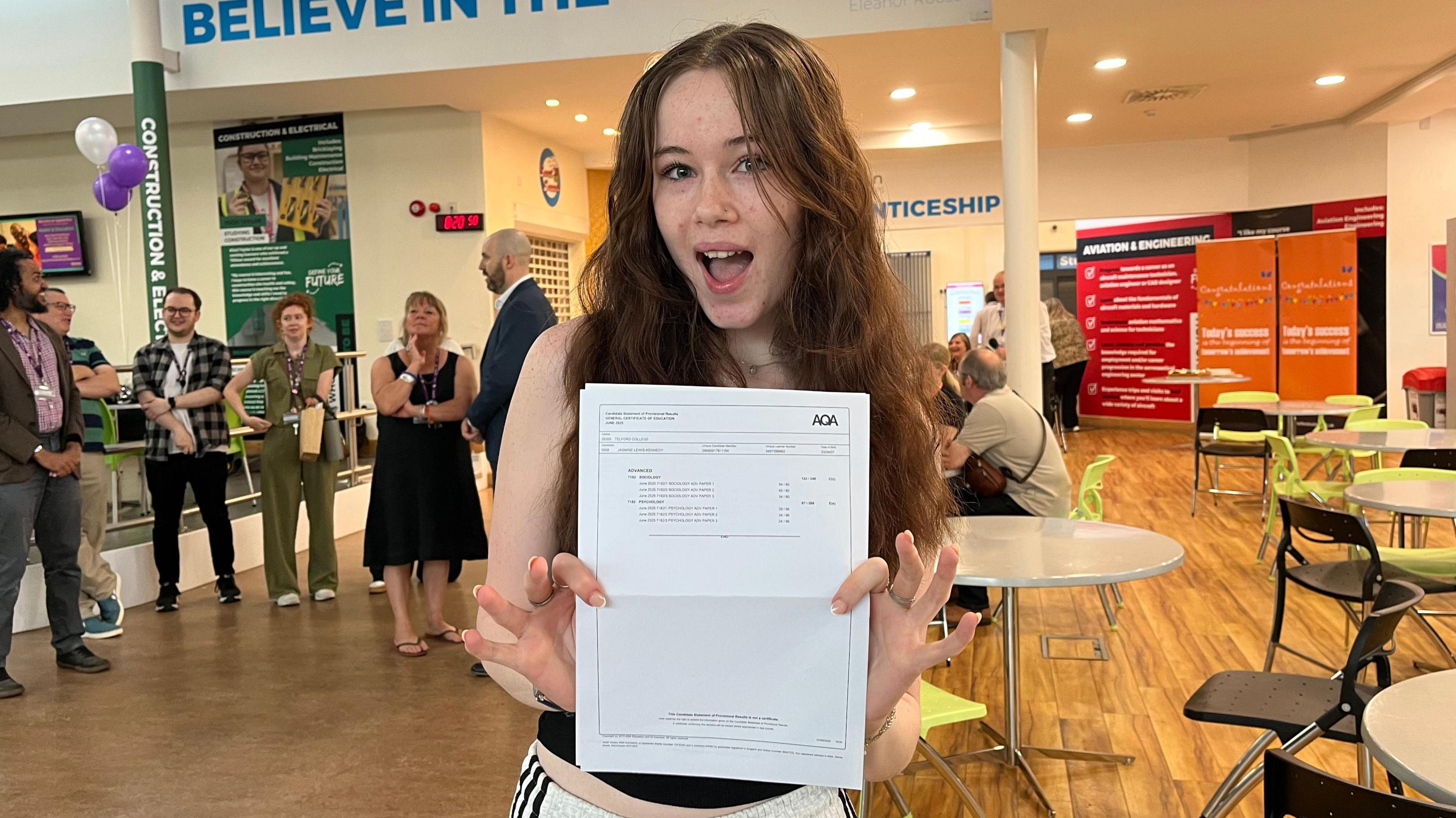 Jasmine is pictured holding up her results - a white piece of paper, with little detail visible apart from 'AQA' written in bold in the top right corner. Jasmine is smiling widely, and is wearing a black vest and grey joggers. She's stood in a school atrium, which has white walls and the word 'Believe' written on them.