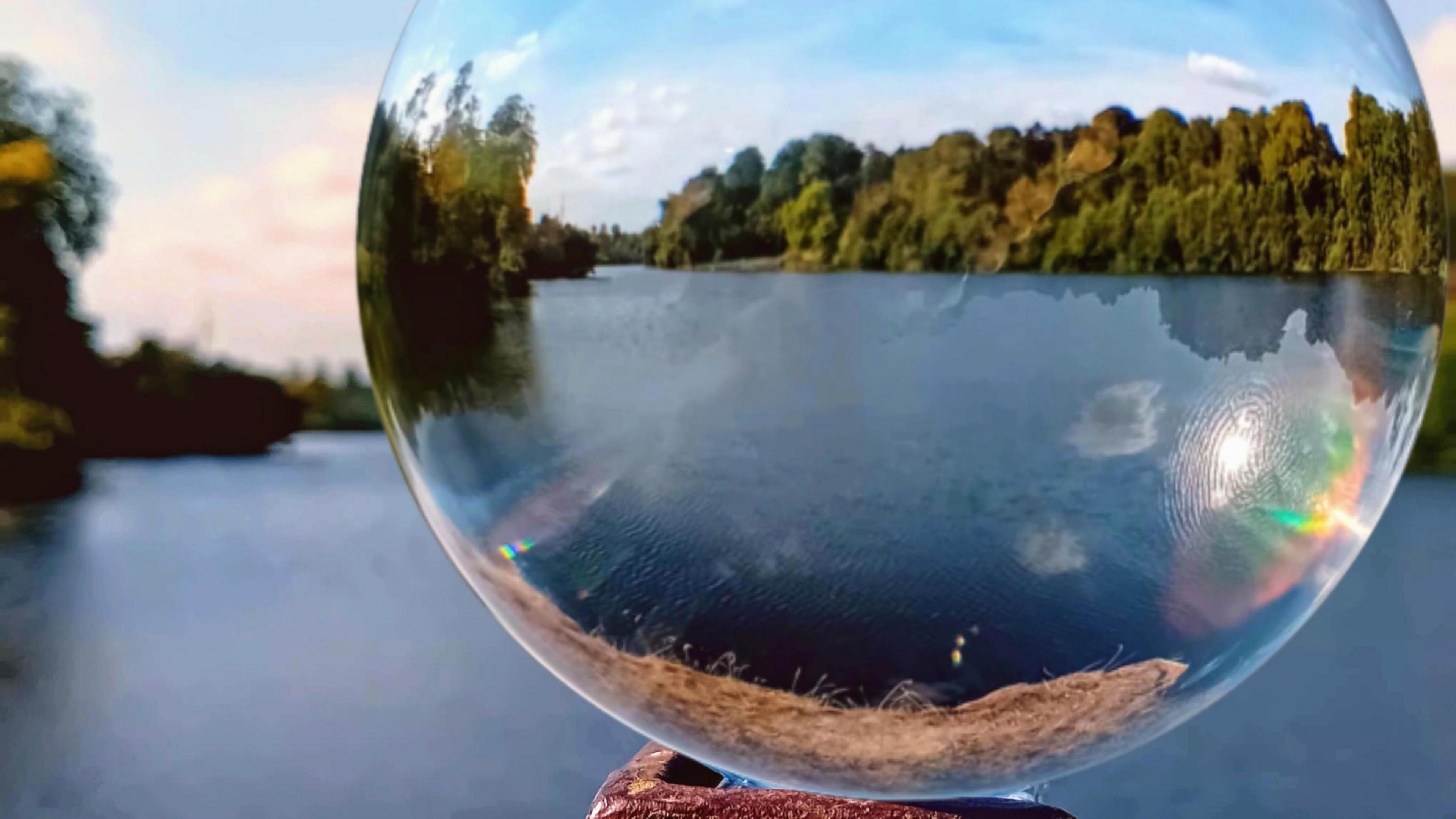 The view of a lake as seen through a glass ball. Trees, water and a blue skyline can be seen in the background, through the ball. The rest of the background is out of focus.