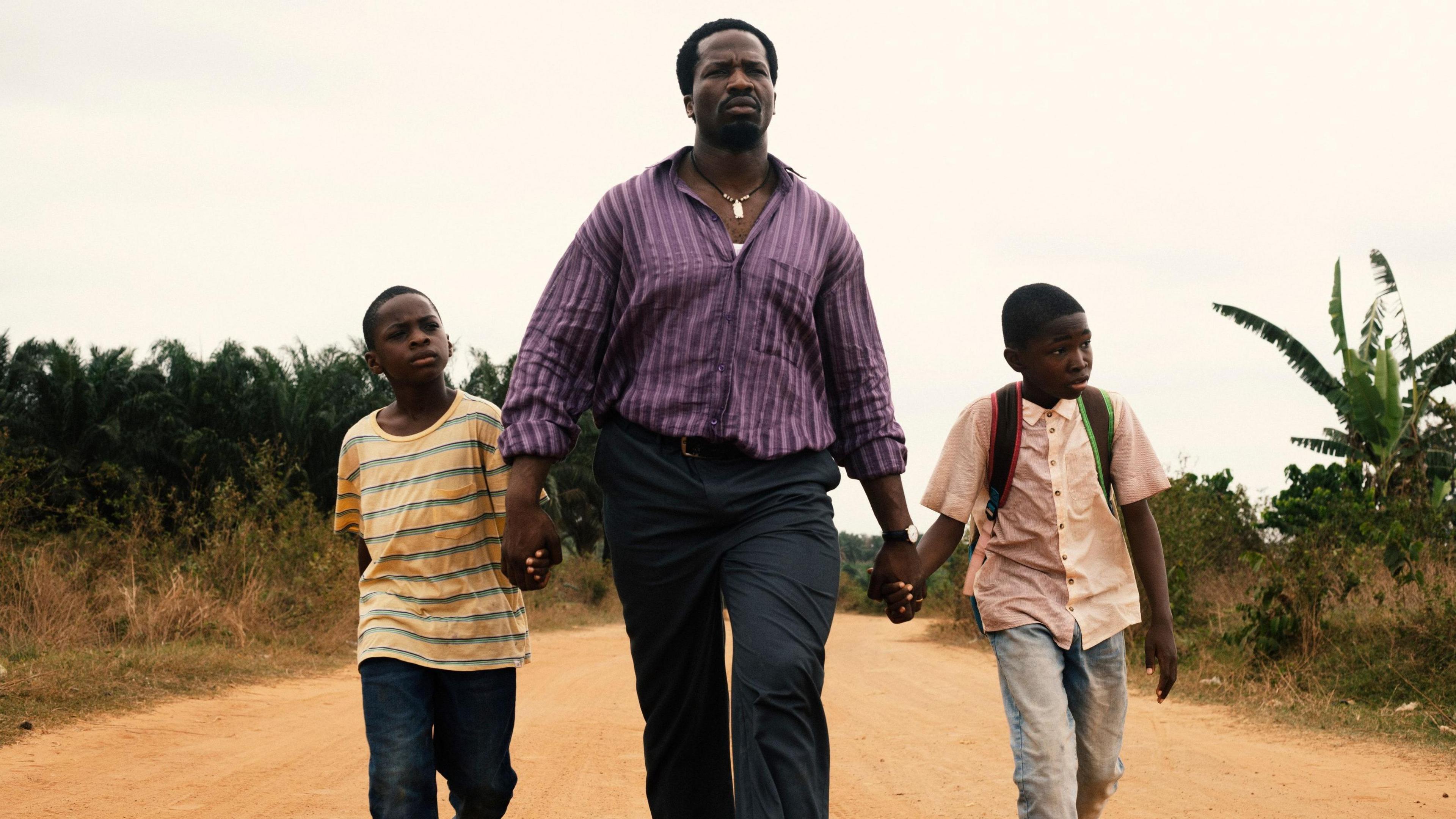 Actor Sope Dirisu walks down a dusty road whole holding the hands of young actors Godwin Chiemerie Egbo and Chibuike Marvellous Egbo. Sope is wearing a purple striped shirt and jeans. The boys are wearing jeans and T-shirts. 