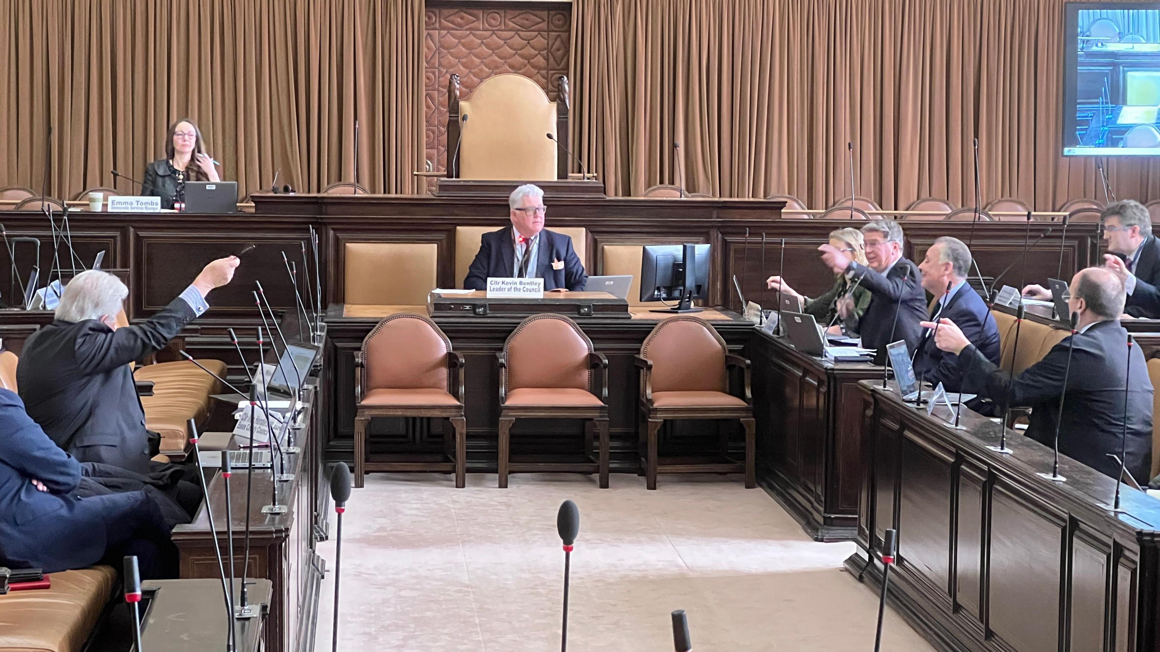 A council chamber with a meeting taking place.  A grey haired man with glasses is sat at the head of the meeting - Kevin Bentley - with two opposing benches. Councillors have their hands up pointing in the air during a debate.