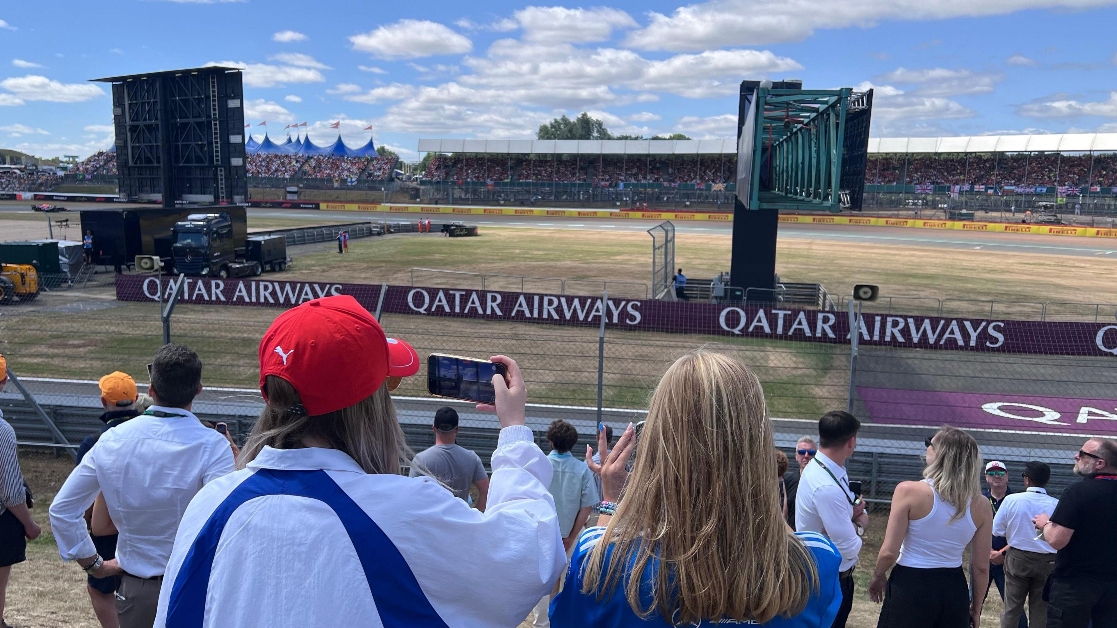 View of fans watching the sprint race at the British Grand Prix. They include a female fan in a red Ferrari hat who is taking a picture on her smartphone. the background shows a view of the Vale corner of the track and its grandstands.