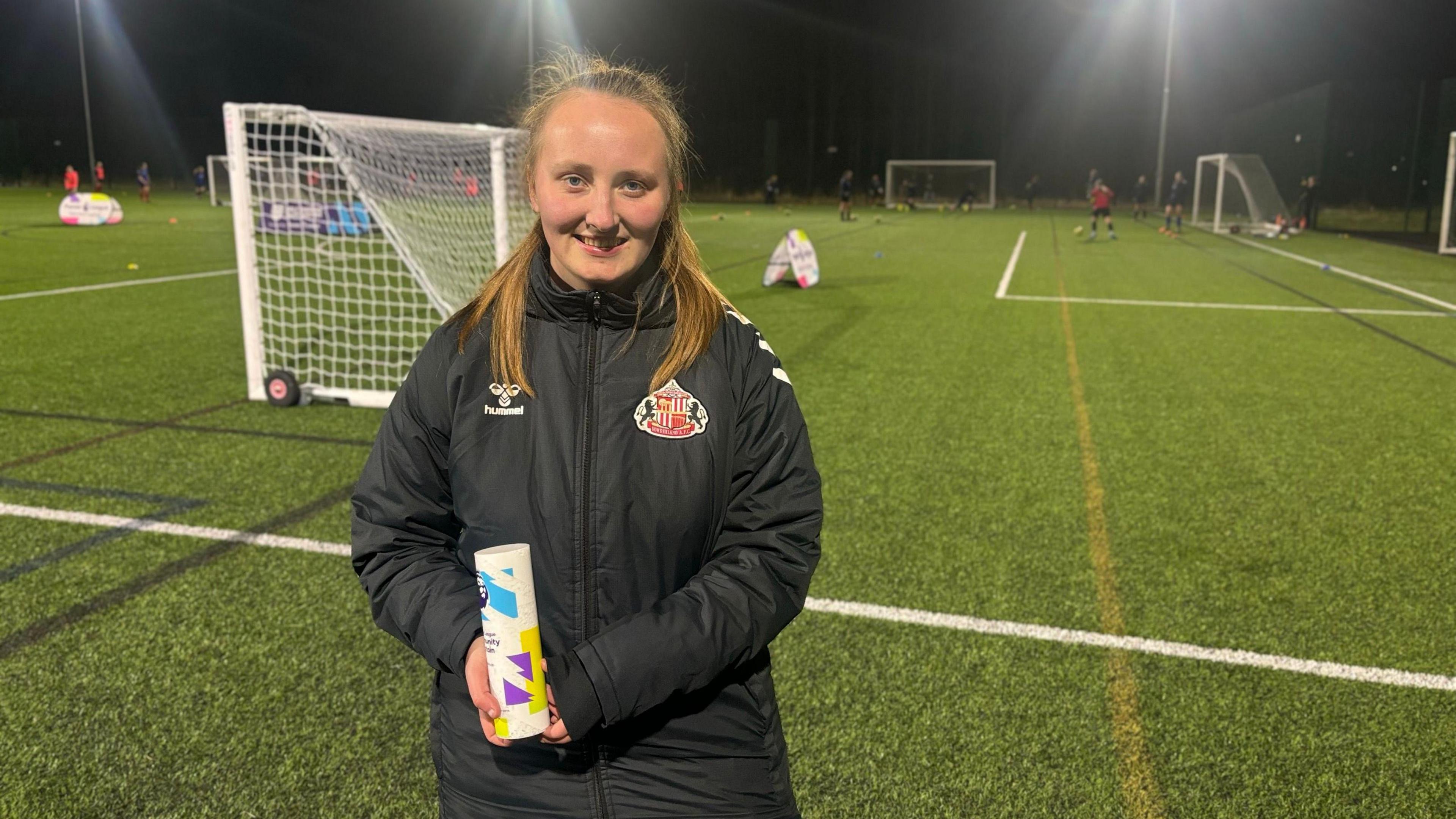 Sophie Gibson who has light hair in a ponytail and is wearing a black coat with the red and white Sunderland AFC badge. She's standing on a football pitch and is holding a white, blue, purple and yellow award.