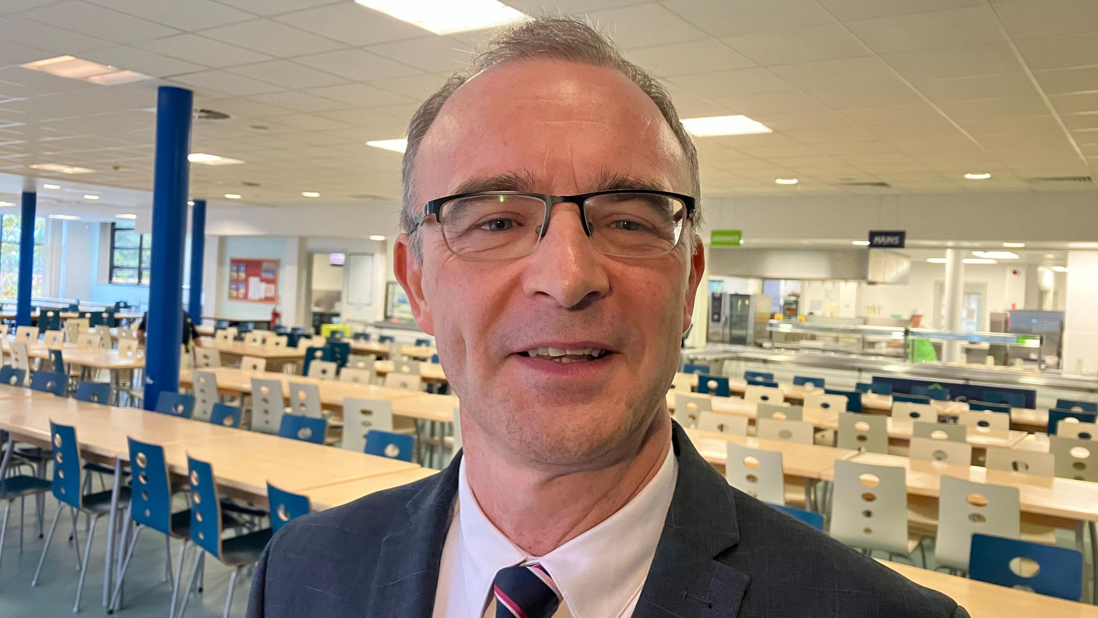 A man standing in a canteen wearing glasses and a suit