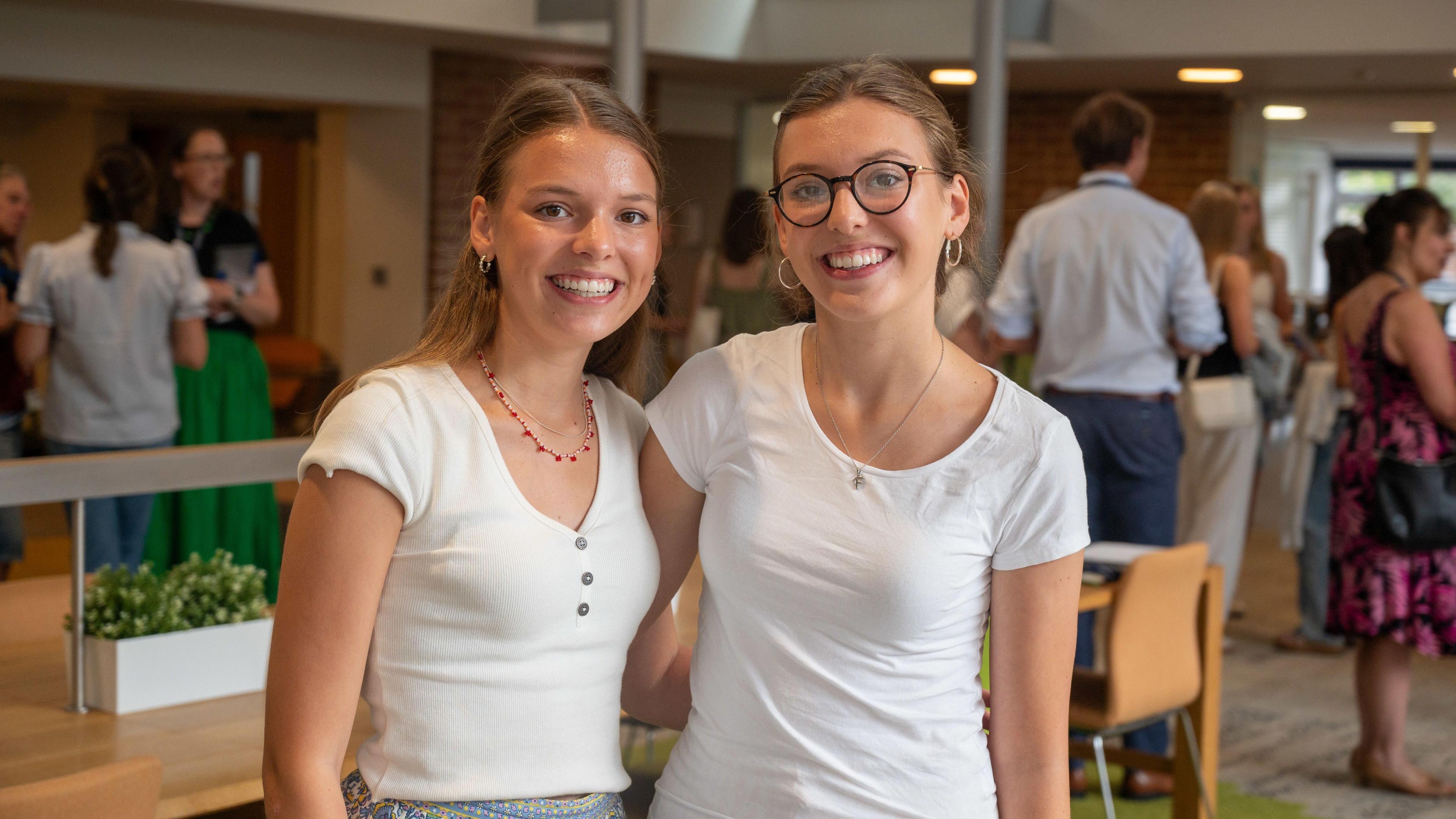 Florence and Alice Bolton stand side by side looking at the camera and grinning widely. They both have dark blonde hair and are wearing white shirt sleeved t shirts. In the background are groups of people gathered inside the college talking.