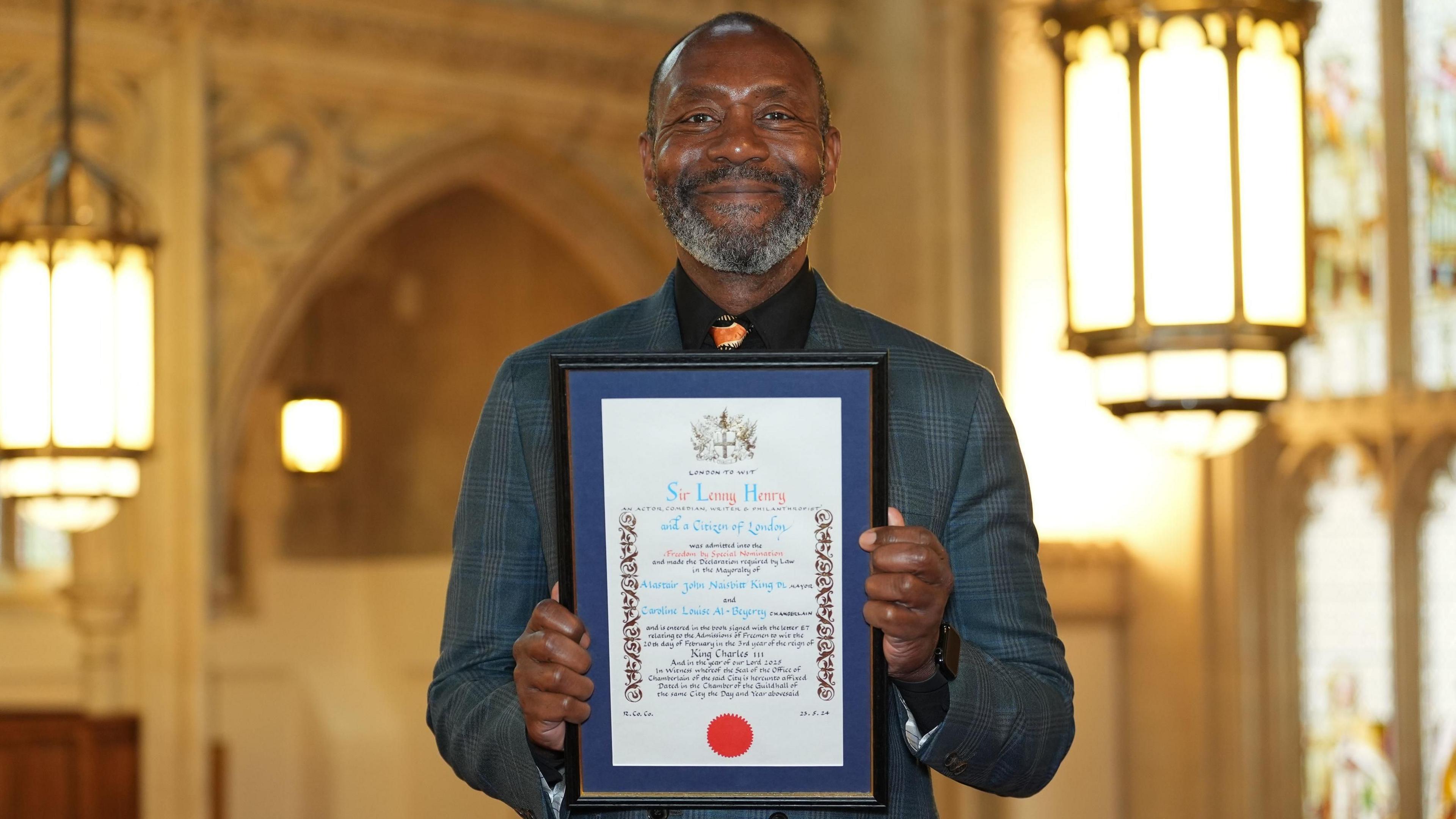 Sir Lenny Henry wearing a grey suit holding certificate showing he had been granted freedom of the City. The interior of the Guildhall is in the background.