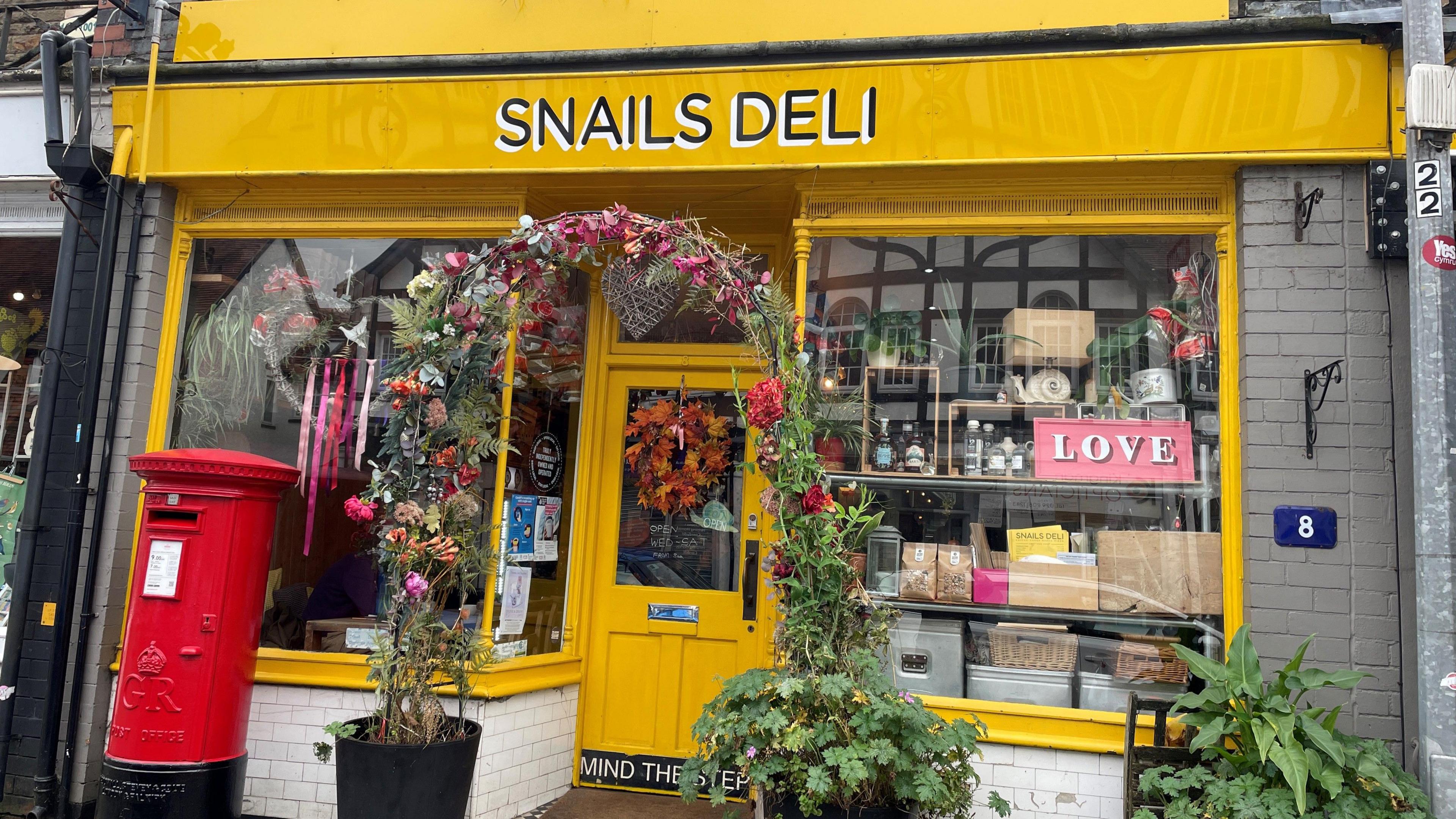 A bright yellow shop front, with a red post box in front of it. There are also various decorative elements including a pink sign that says love and pink tassles on the inside of the shop window. There is also a plant based archway outside. 