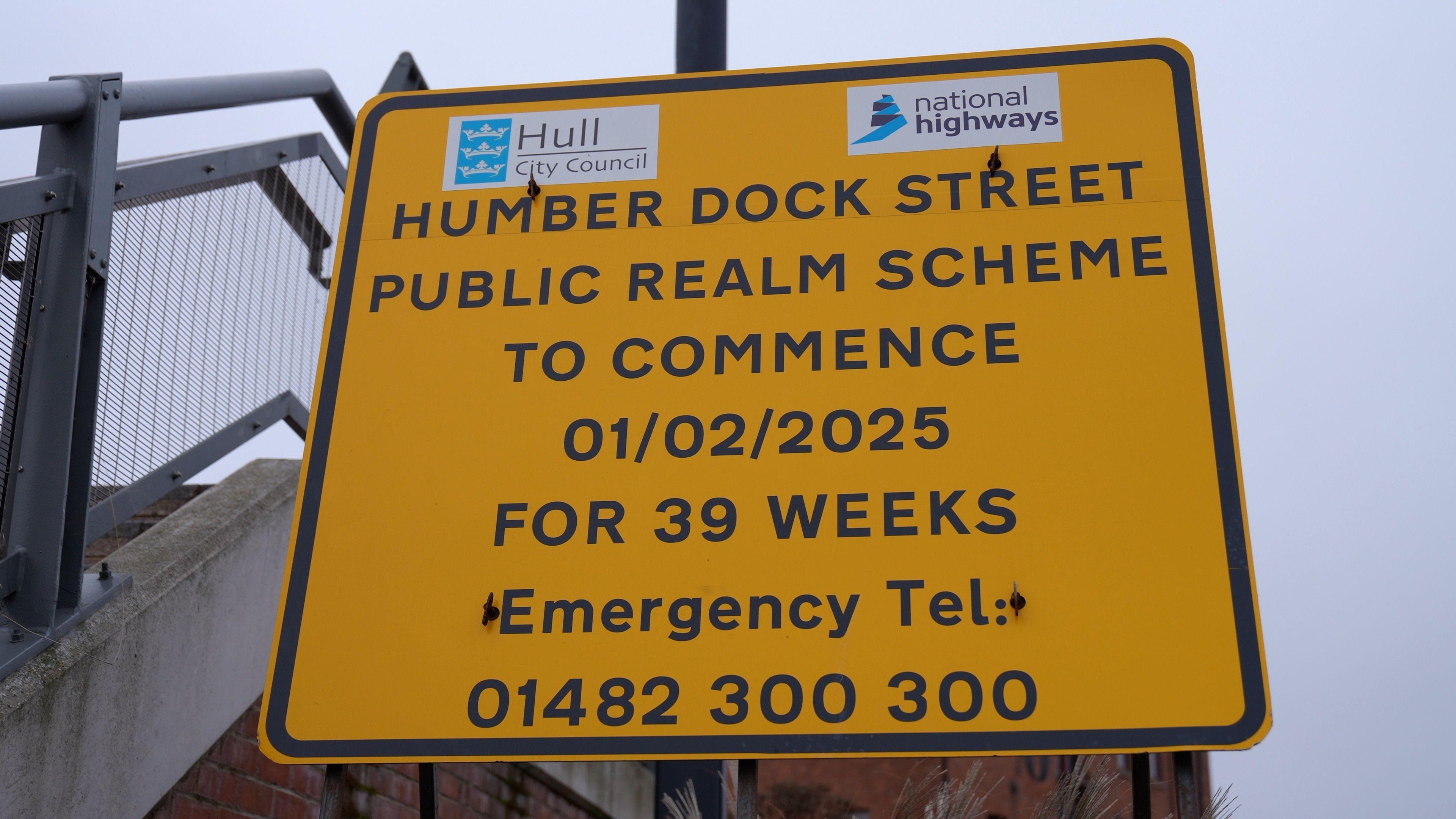 A bright yellow roadside sign with black text is mounted on a metal post near a railing. At the top, there are two logos: one for "Hull City Council" on the left and one for "National Highways" on the right. The sign is outdoors, with a grey overcast sky in the background and part of a metal fence visible on the left. The main text reads: "HUMBER DOCK STREET PUBLIC REALM SCHEME TO COMMENCE 01/02/2025 FOR 39 WEEKS. Emergency Tel: 01482 300 300.”