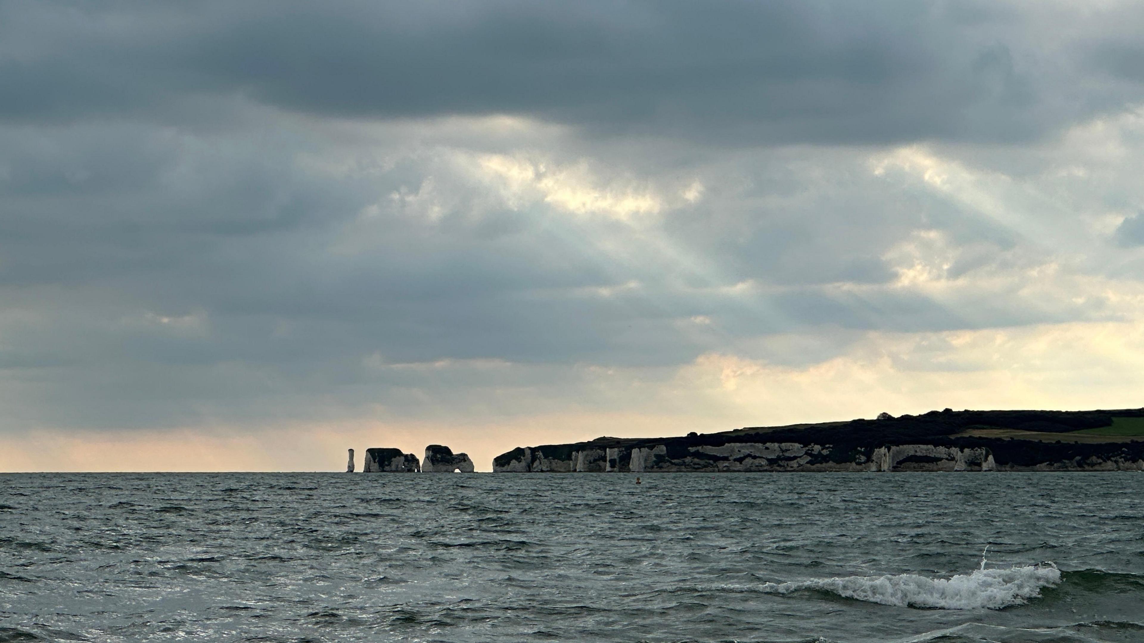 A coastal shot of the sea with cliffs in the background. The sky is grey with thick clouds but rays of sun can be seen coming through a few gaps