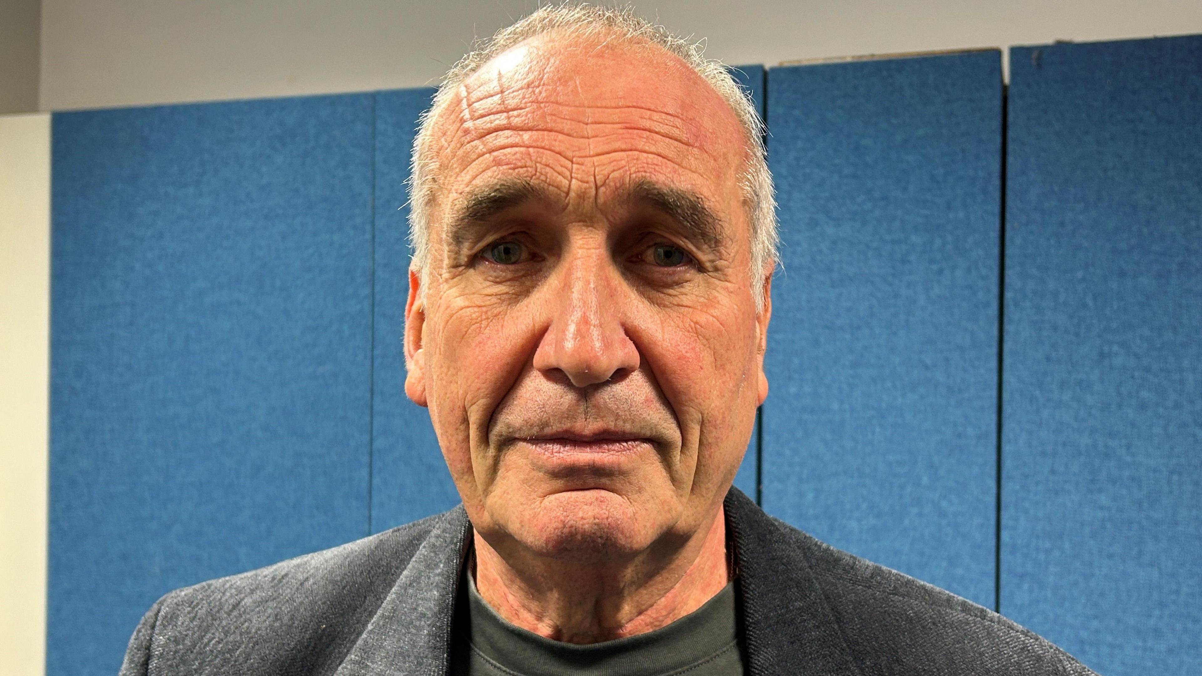 A man with grey hair and a dark grey jacket stood in front of a blue background.