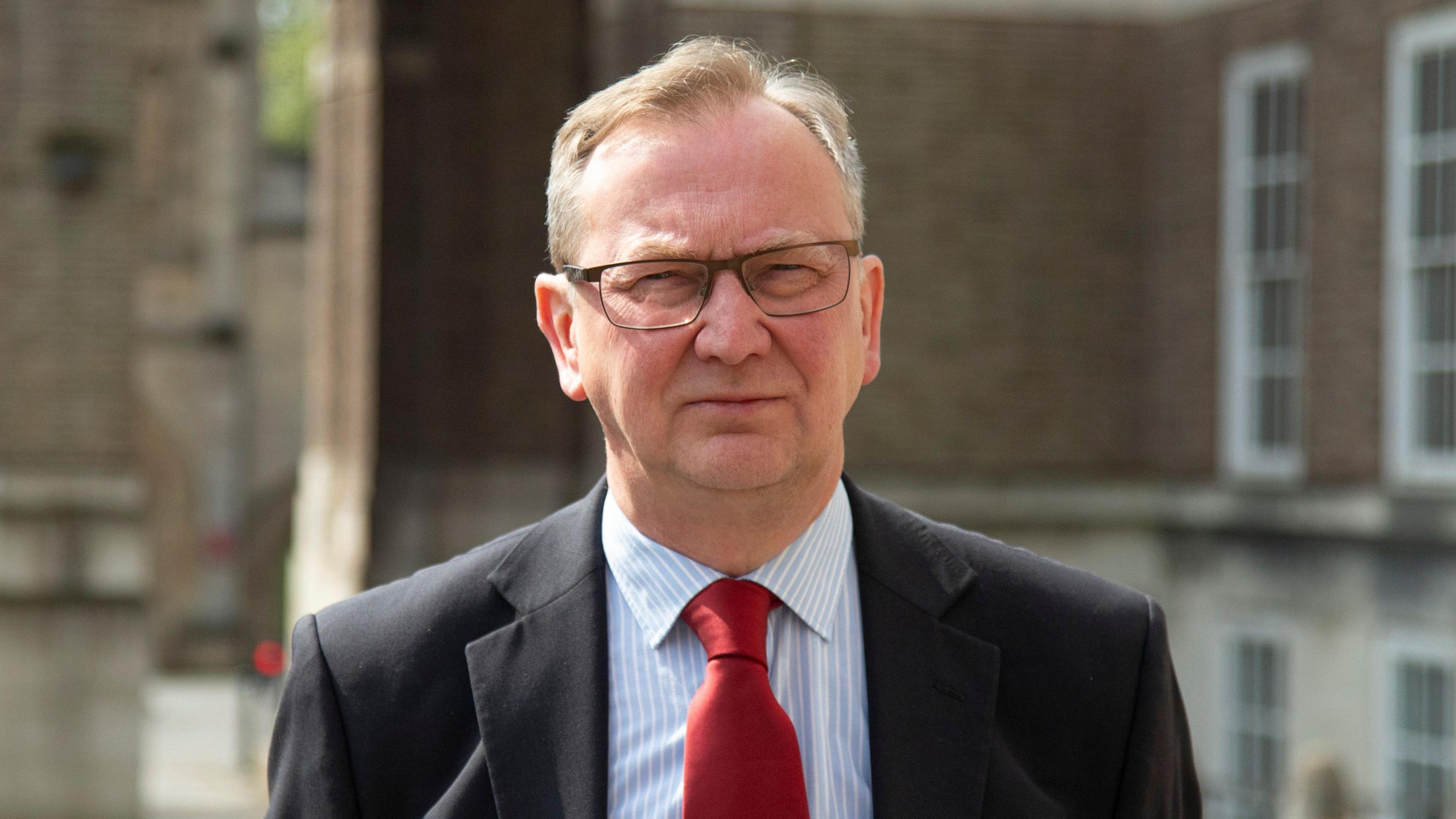 A head and shoulders shot of councillor Fabian Breckels, who is is squinting as he looks at the camera. He has short grey hair and wears rectangular, thin-rimmed glasses. He is wearing a black suit jacket, white and blue pin-striped shirt and a red tie. The council building is blurred behind him.