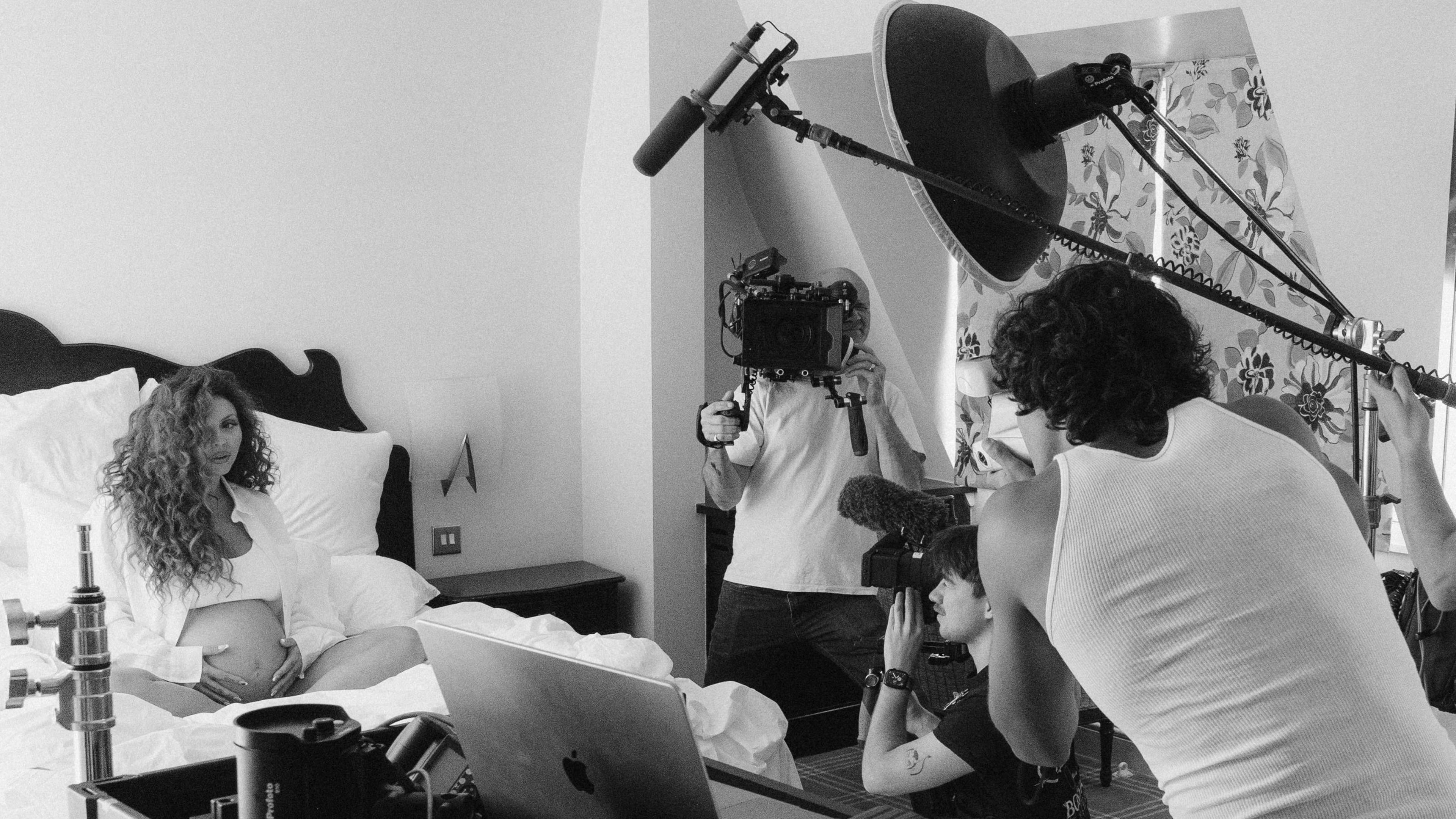 A black and white photo of Jesy Nelson sitting on a bed holding her baby bumb. There is film crew surrounding her with cameras and sound booms. 