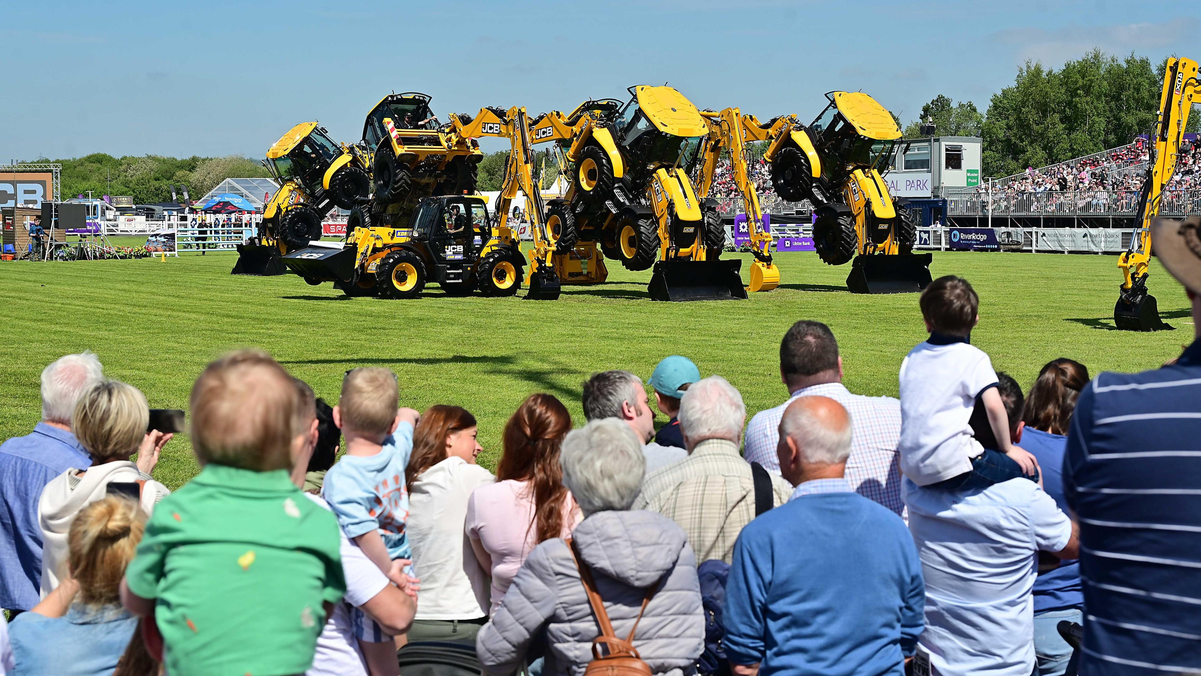 Crowds watching yellow JCB diggers in a field.